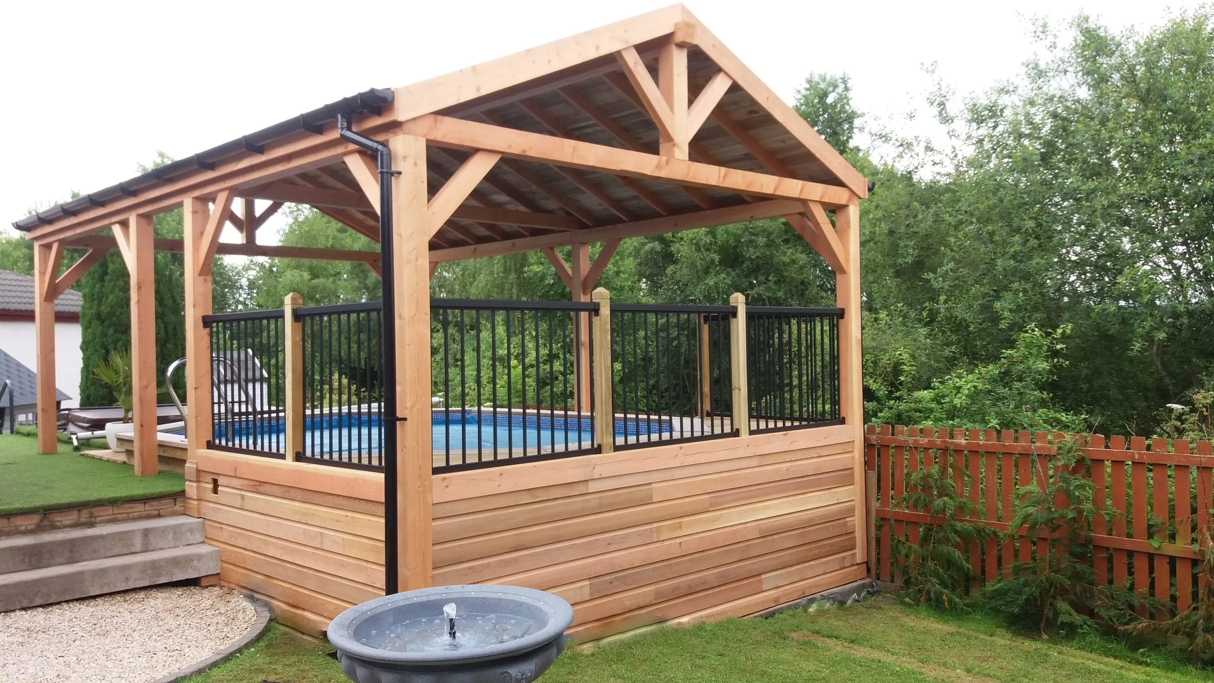 A wooden gazebo with a sloped roof and black metal railing surrounds an above-ground swimming pool, located in a backyard with green trees and a wooden fence.