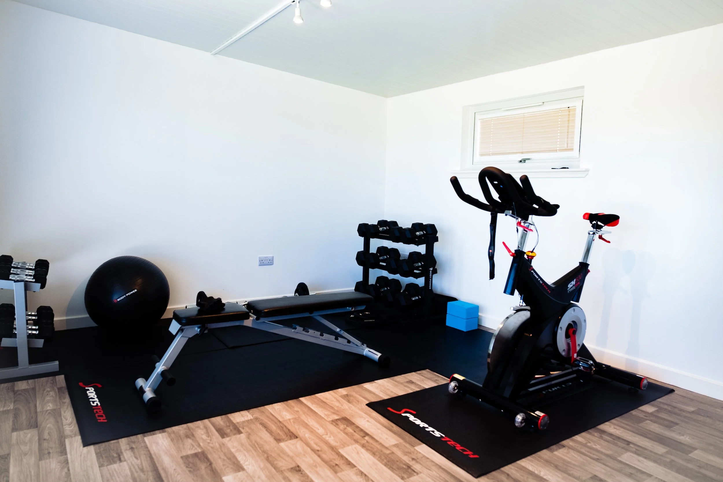 Home gym with exercise bike, weight bench, dumbbells, stability ball, and small blue storage block on a black rubber mat in a room with wood flooring and a small window.