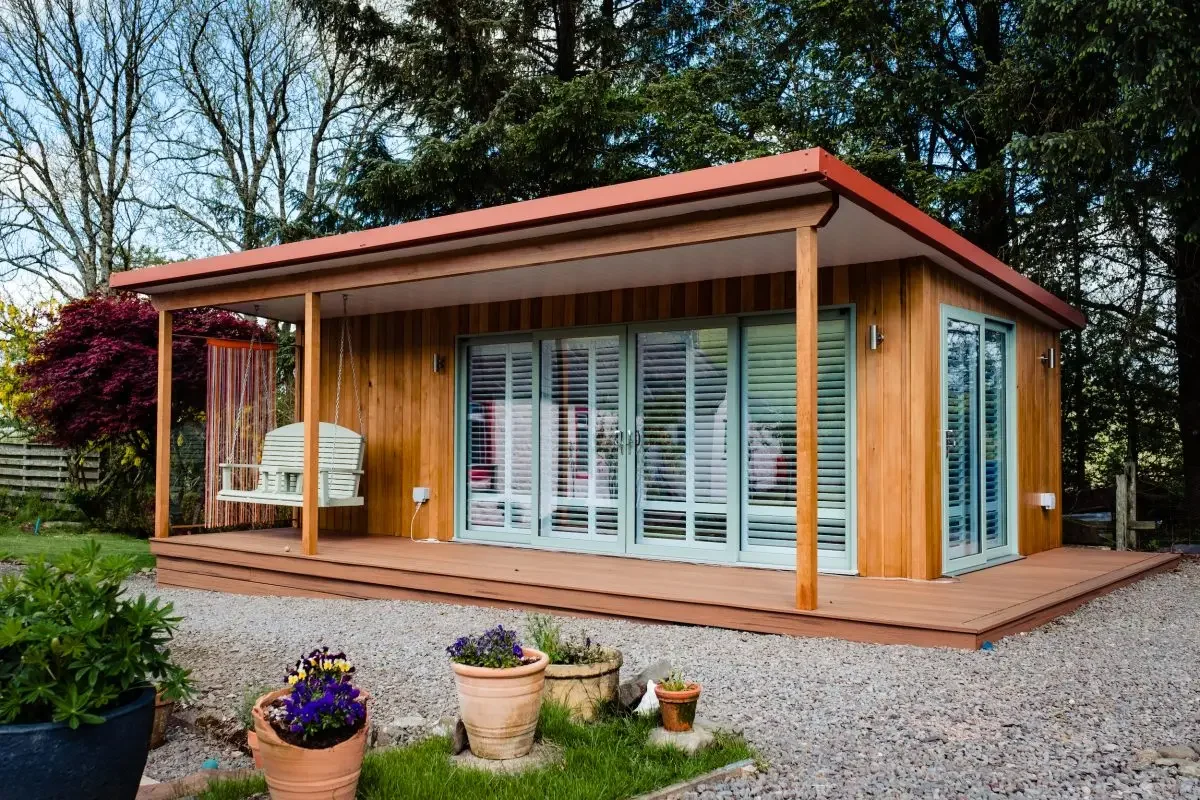 A modern wooden house with a spacious deck featuring a hanging swing chair and potted plants in the garden, surrounded by trees.