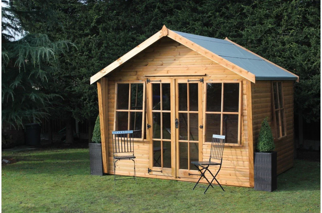 A small wooden garden shed with large windows, a metal roof, and two black metal chairs outside, surrounded by greenery.