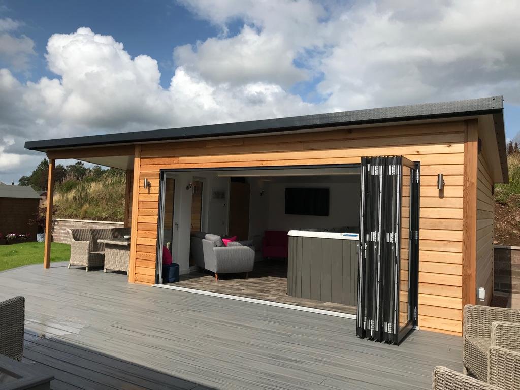 Outdoor wooden deck with a modern tiny house or studio with open sliding glass doors showing a living room with sofas, a hot tub, and outdoor furniture under a cloudy sky.