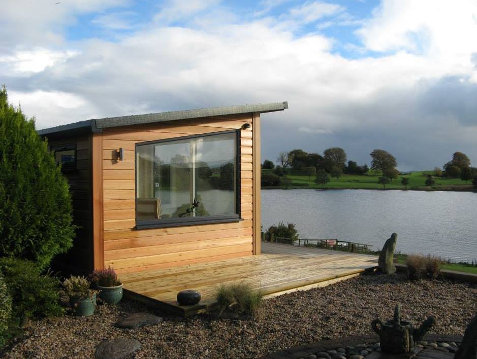 Small wooden cabin by a lake with a deck, surrounded by plants and rocks, under a cloudy sky.