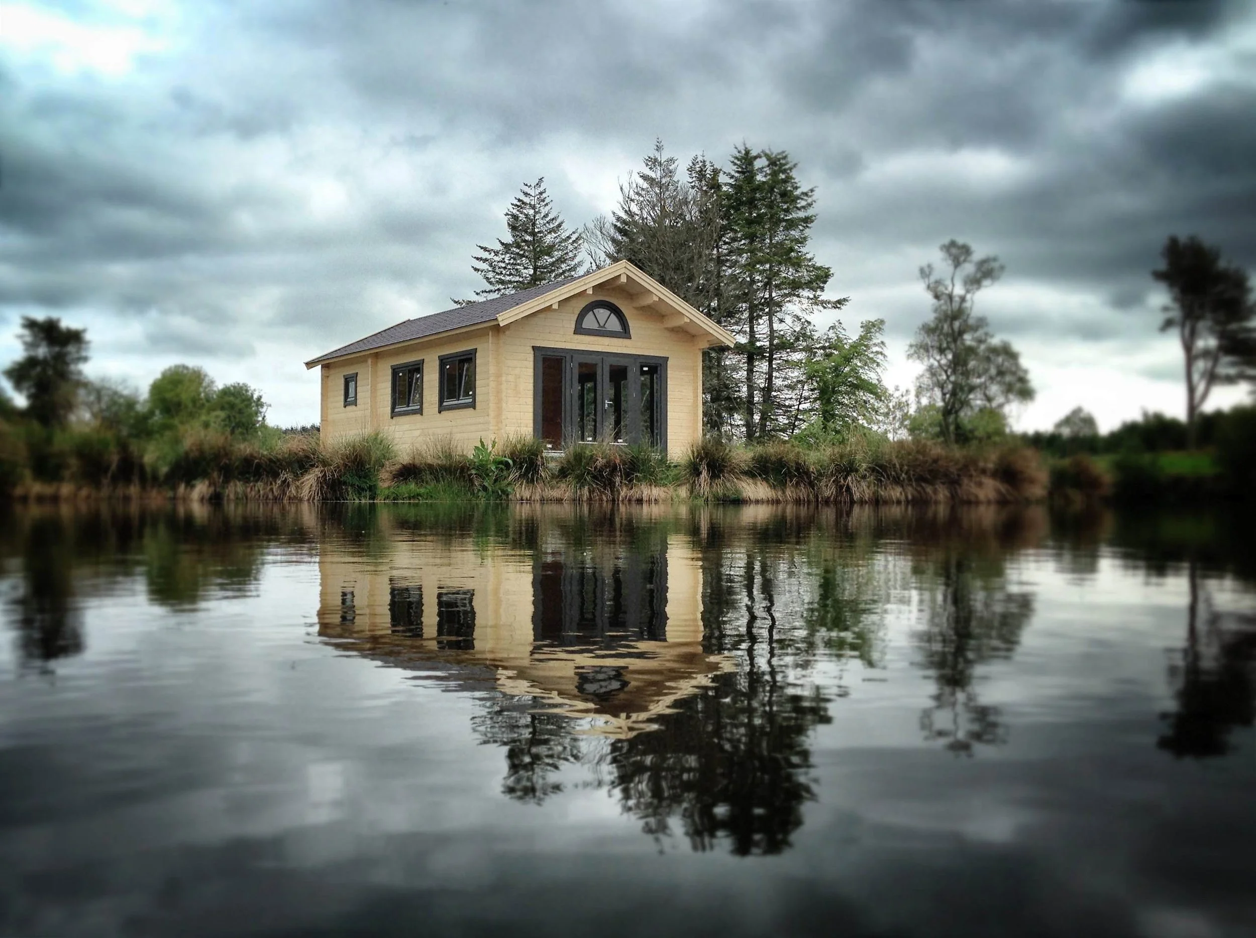 A small wooden house with large windows and a glass doorway is situated on the edge of a body of water, with trees in the background and a cloudy sky overhead, and the house is reflected in the water.
