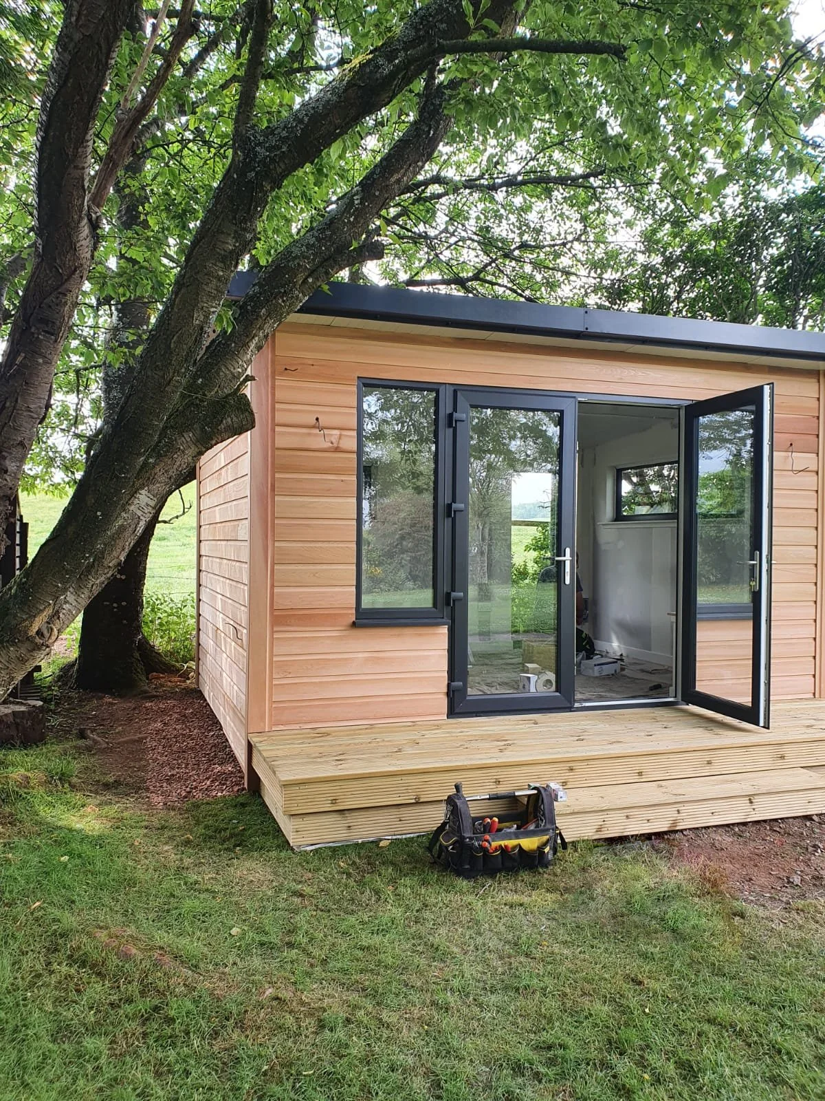 A small wooden shed with black-framed glass doors and windows, built on a wooden deck, situated in a backyard with grass and a large tree nearby.
