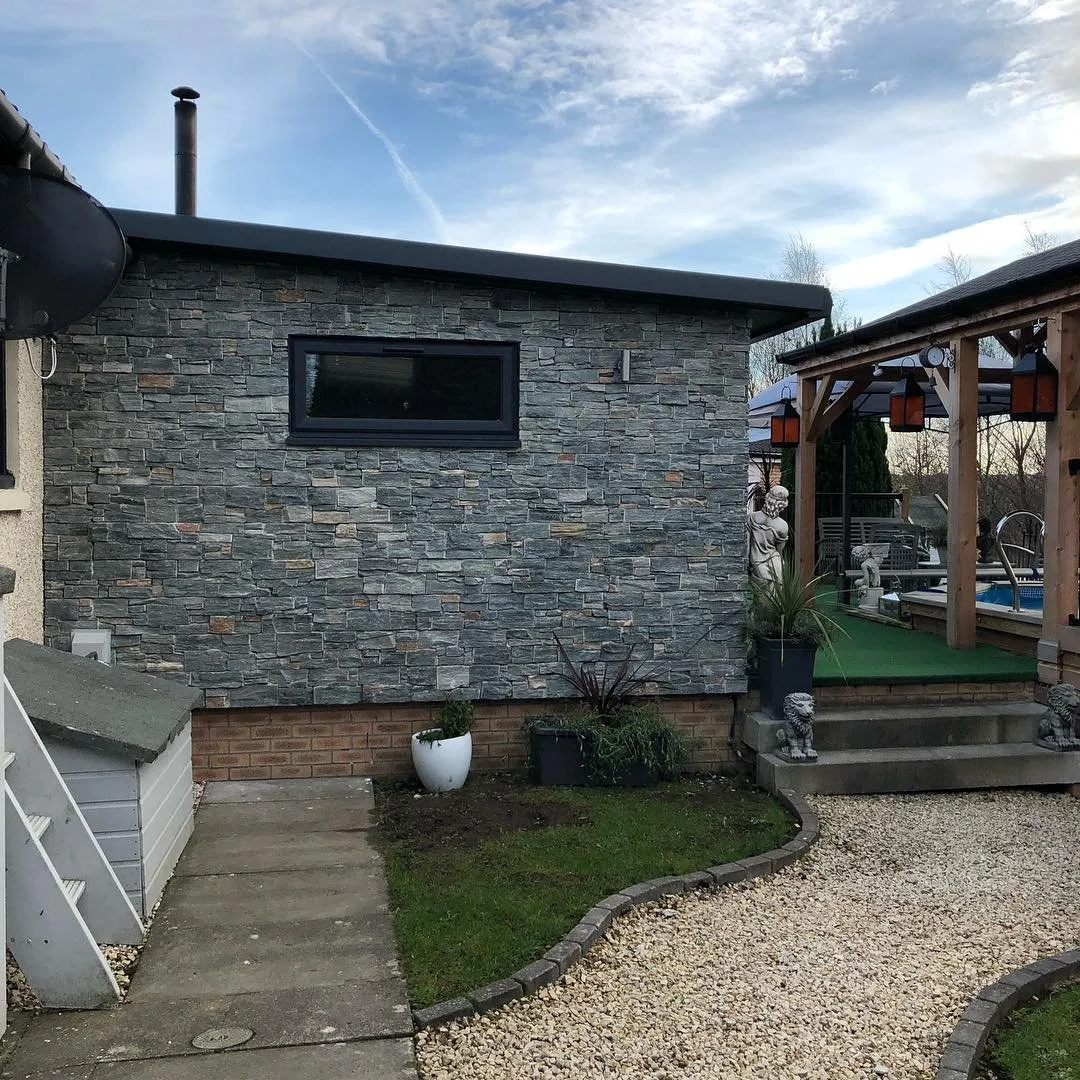 A backyard scene with a stone wall, a raised deck with steps, decorative statues, potted plants, and a gravel pathway, under a blue sky with some clouds.