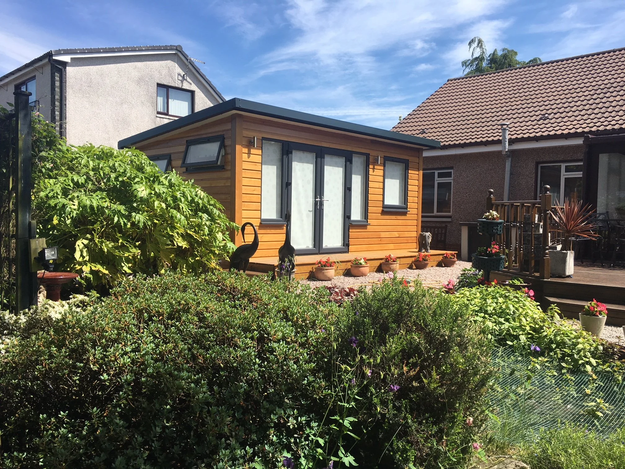 A backyard garden with a small wooden house, surrounded by green plants, shrubs, and colorful potted flowers, under a blue sky with some clouds.