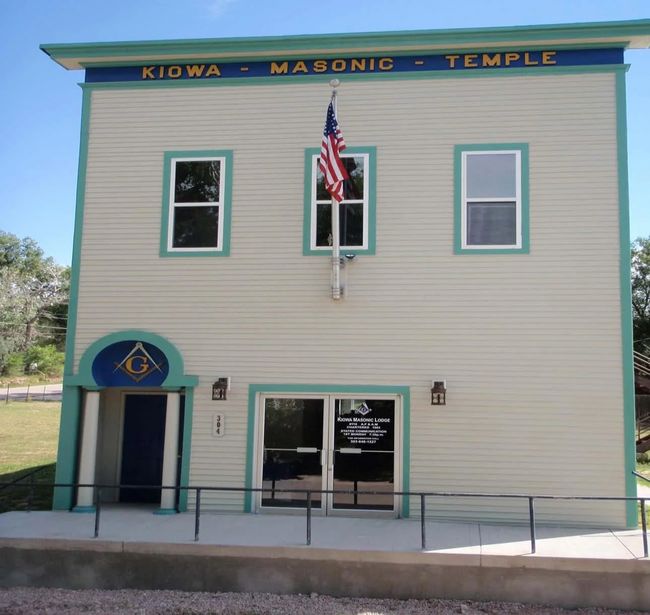 A two-story building painted in light beige with teal trim, labeled as Kiowa Masonic Lodge. It has three windows on the upper floor, a small arched entrance with a blue sign featuring the Masonic square and compass symbol, and a glass door with a sign displaying the lodge's name and contact information. An American flag is mounted on a pole in the center of the building's facade.