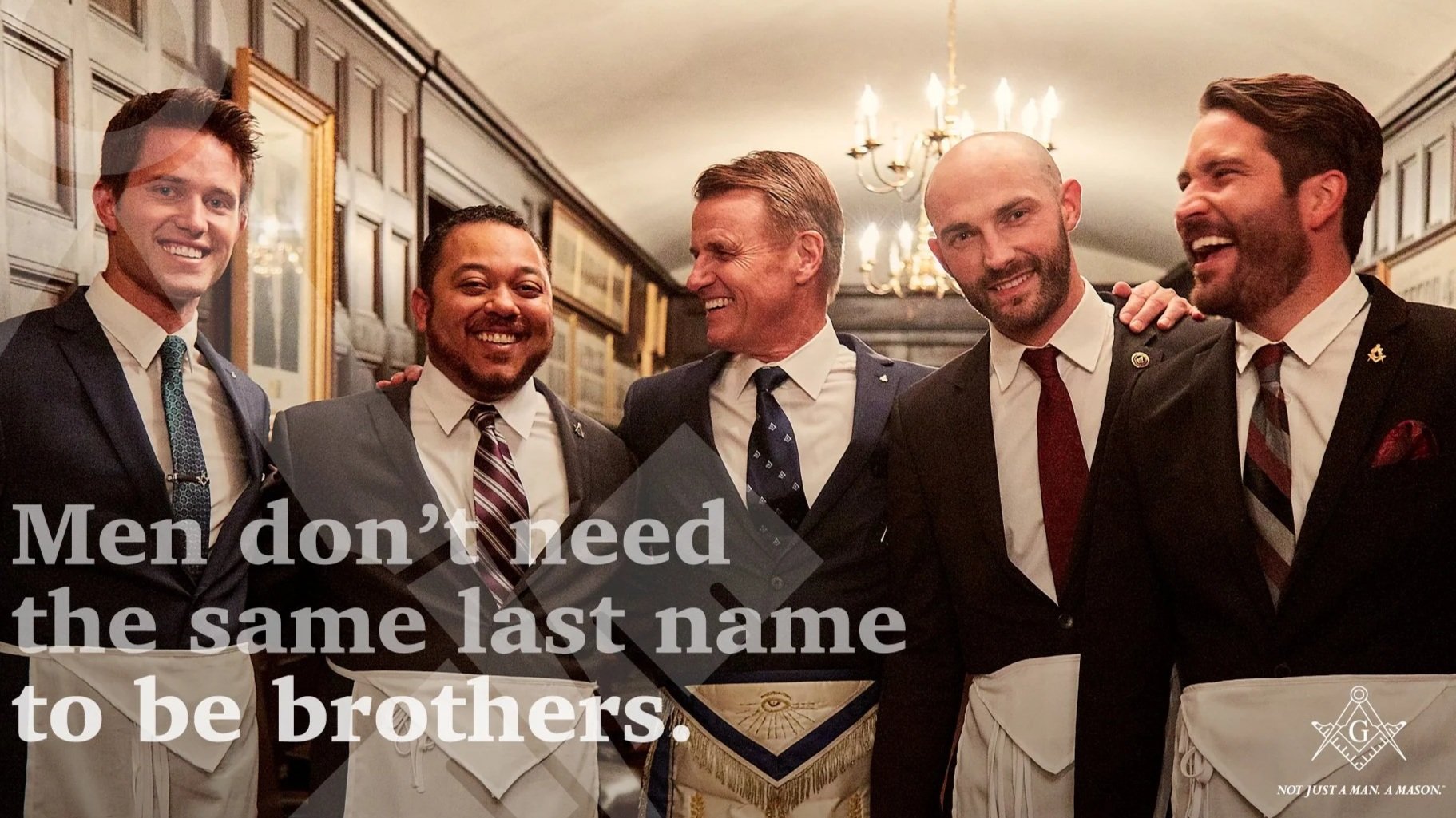 Group of men in formal suits and aprons, smiling and laughing, standing in a decorated room with framed pictures and chandeliers