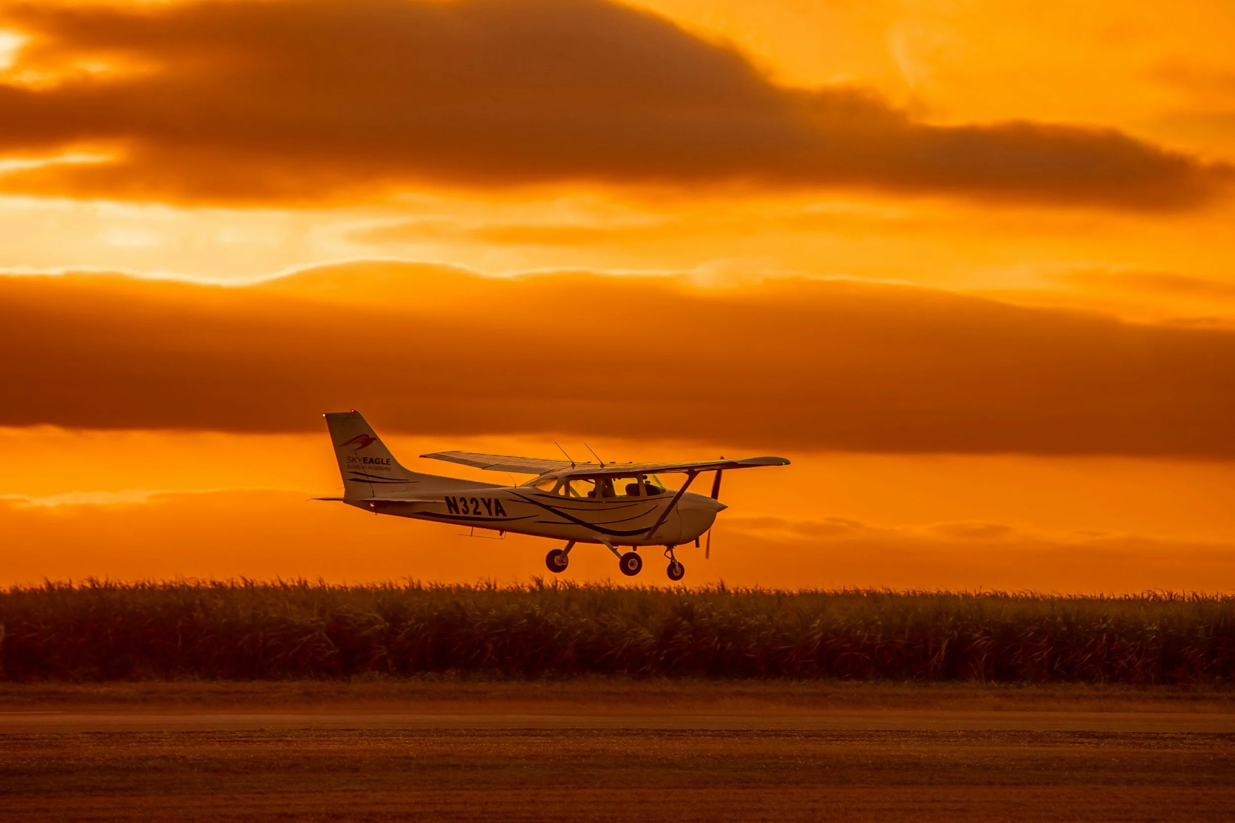 Small airplane flying low over a grassy field during a sunset with orange and yellow sky.