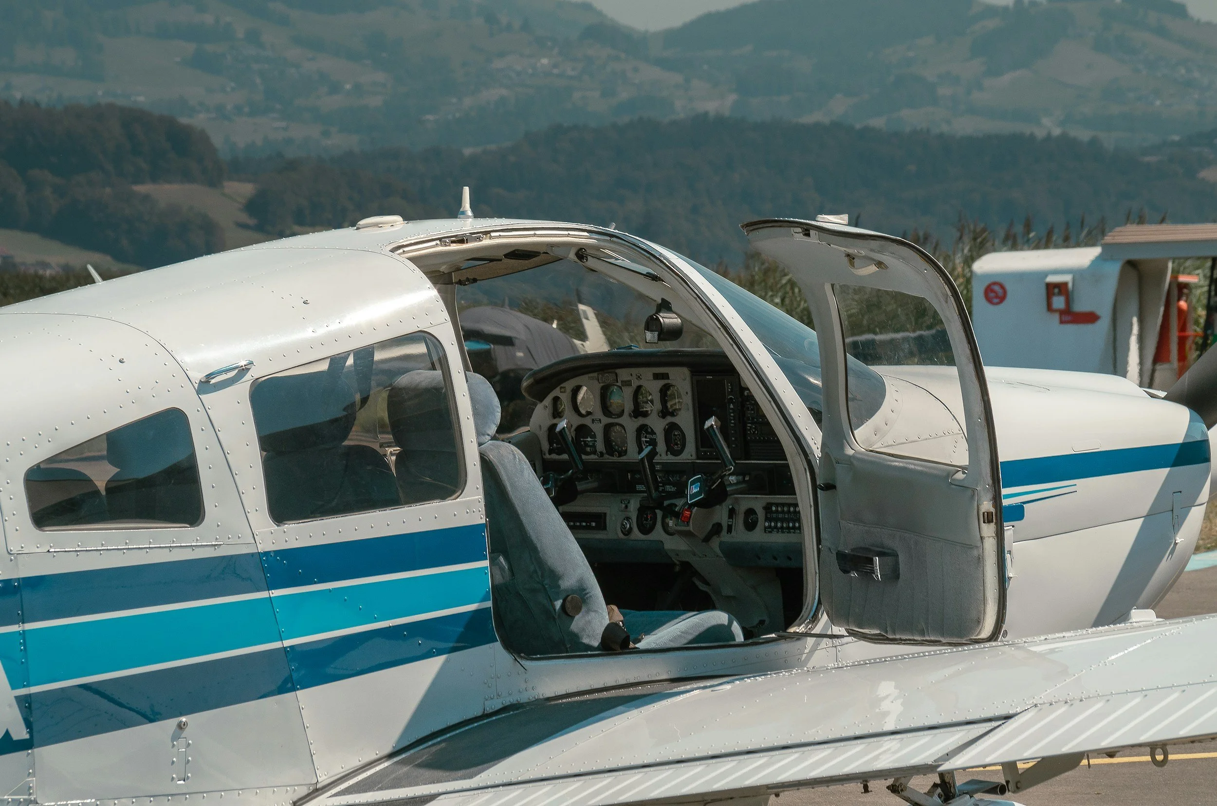 Interior and exterior of a small white and blue airplane on the ground with open cockpit door, showing flight instruments and fabric seats, with a scenic landscape in the background.