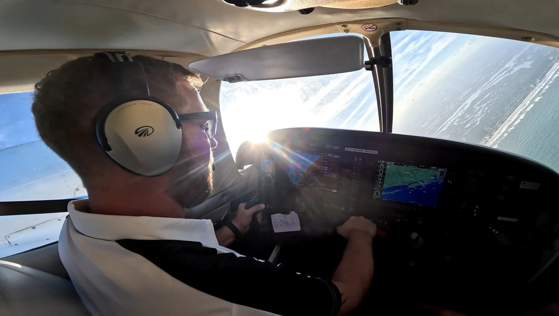 A pilot wearing noise-canceling headphones and sunglasses flying an aircraft during sunset over water, with the instrument panel visible.