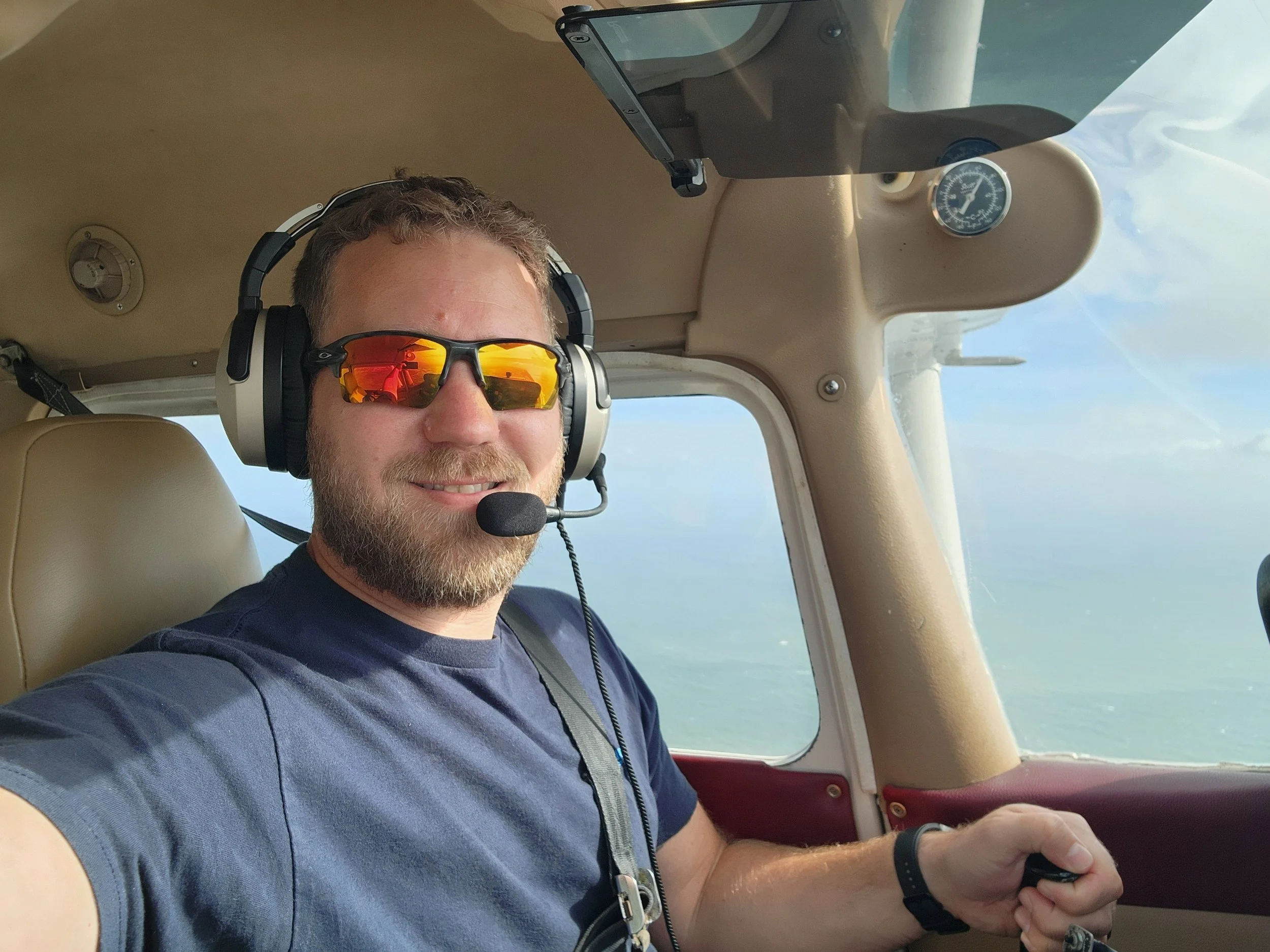A smiling man wearing sunglasses, a headset with a microphone, and a casual blue t-shirt, flying a small airplane over a blue sky with clouds.