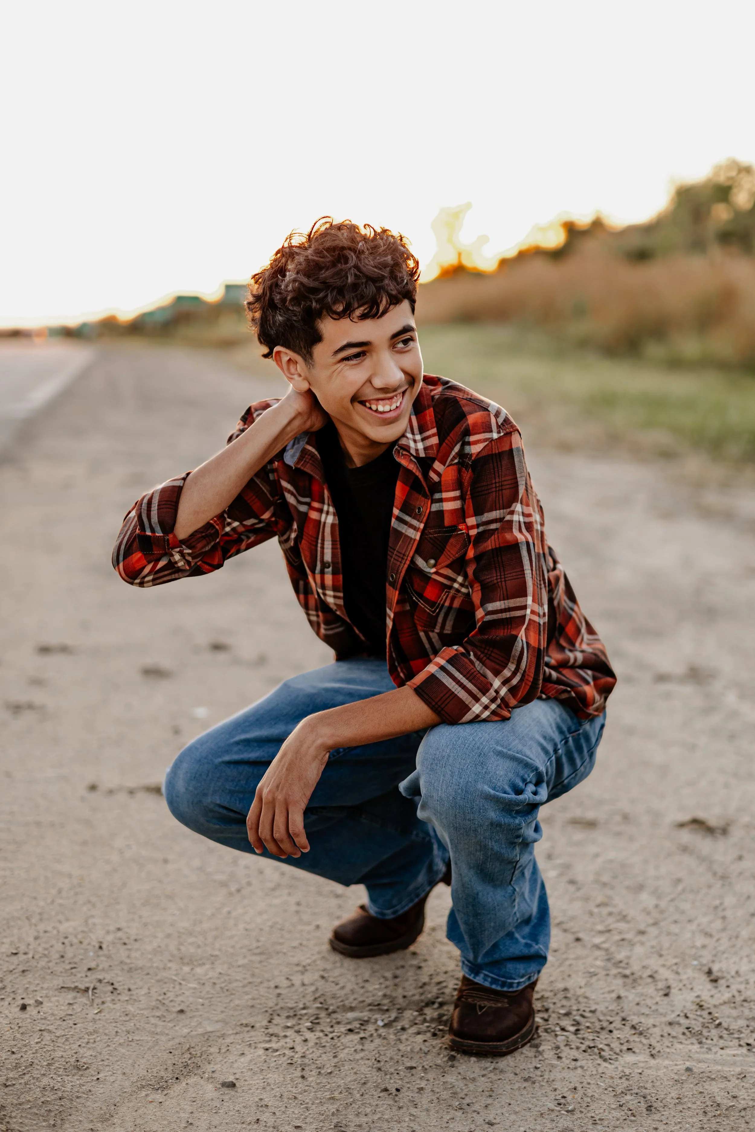 A young man in a red plaid shirt, jeans, and brown boots crouches on a dirt road during sunset, smiling and looking to the side.