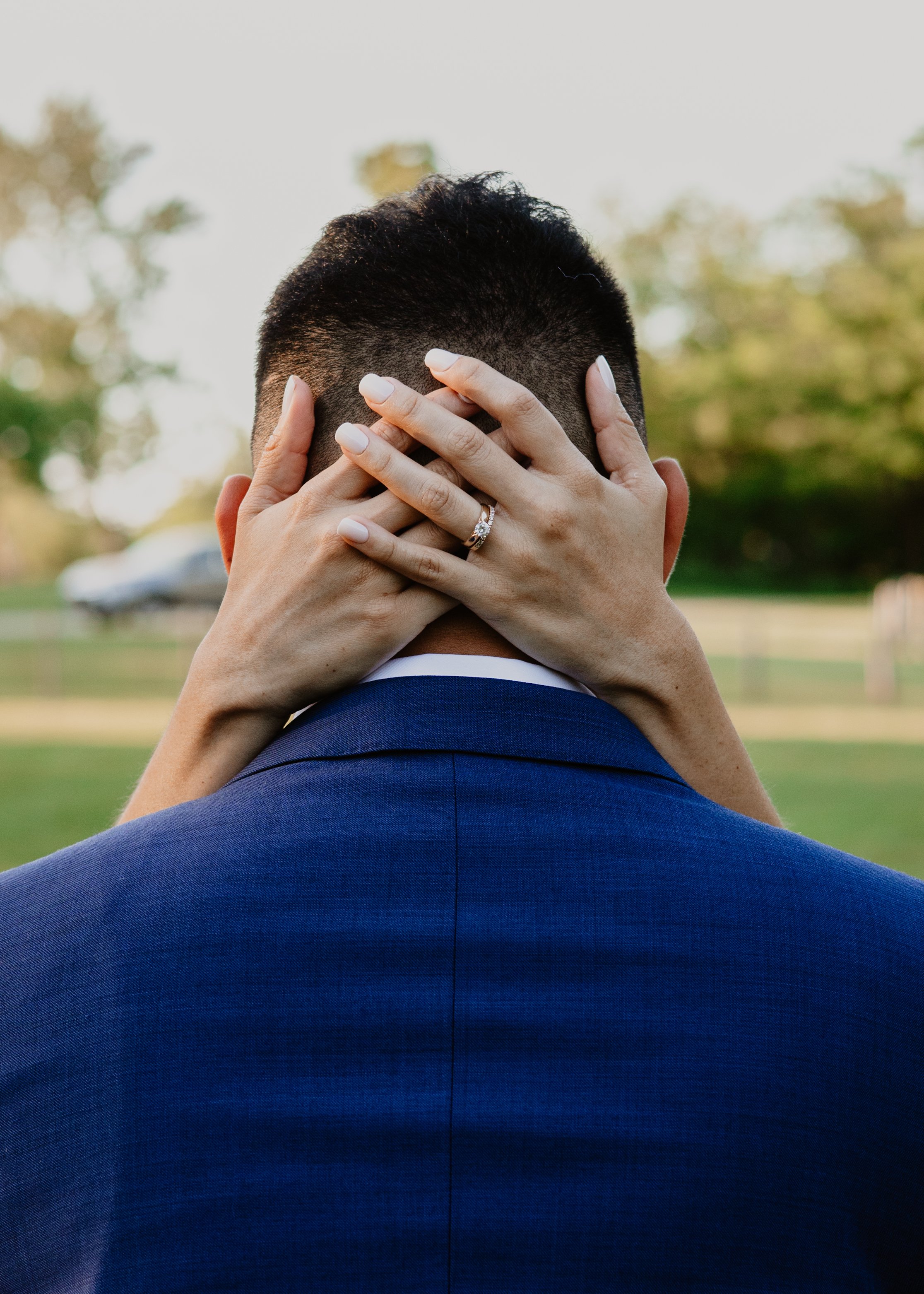 Close-up of a woman holding her hands over a man's face, revealing an engagement ring, outdoors with trees in the background.