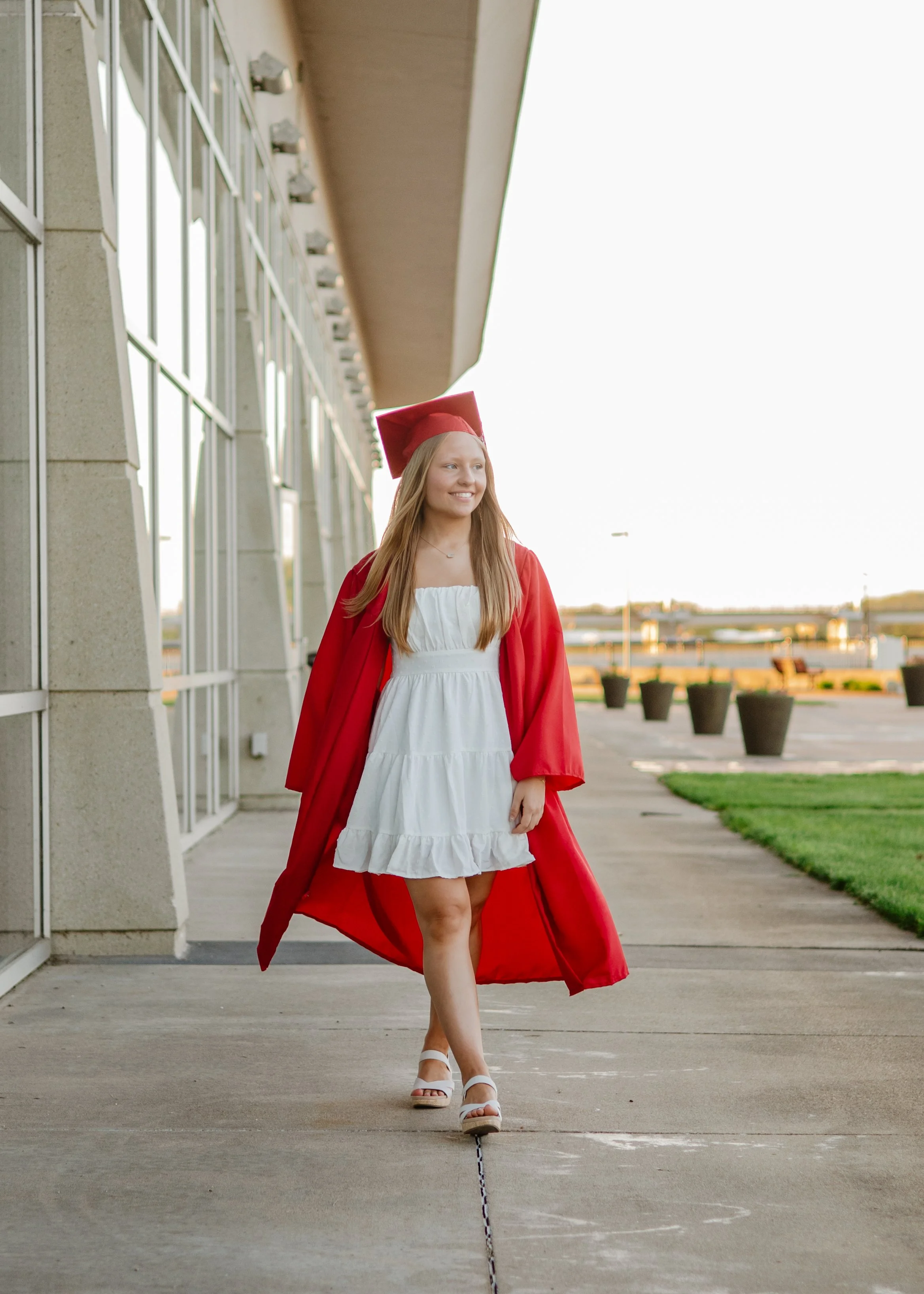 A young woman in a white dress and white sandals wearing a red graduation gown and cap walking outside near a modern building.