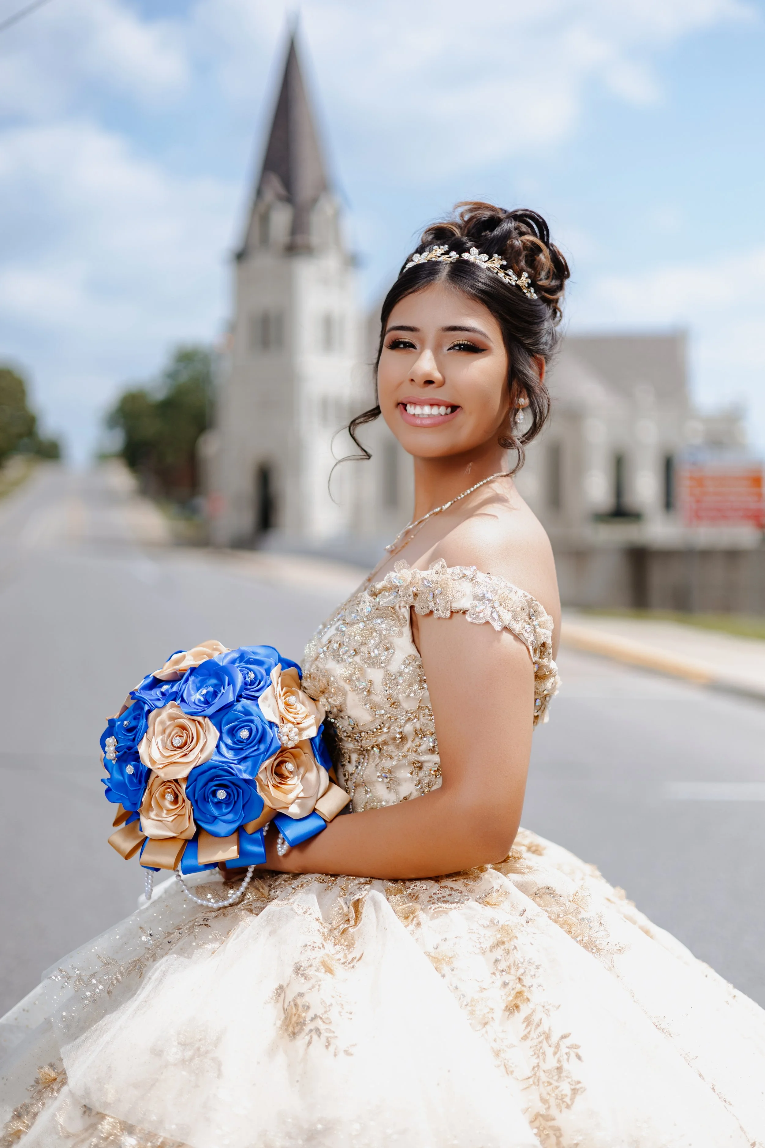 A bride in a wedding dress holding a bouquet of blue and beige roses, standing outdoors with a church steeple in the background.