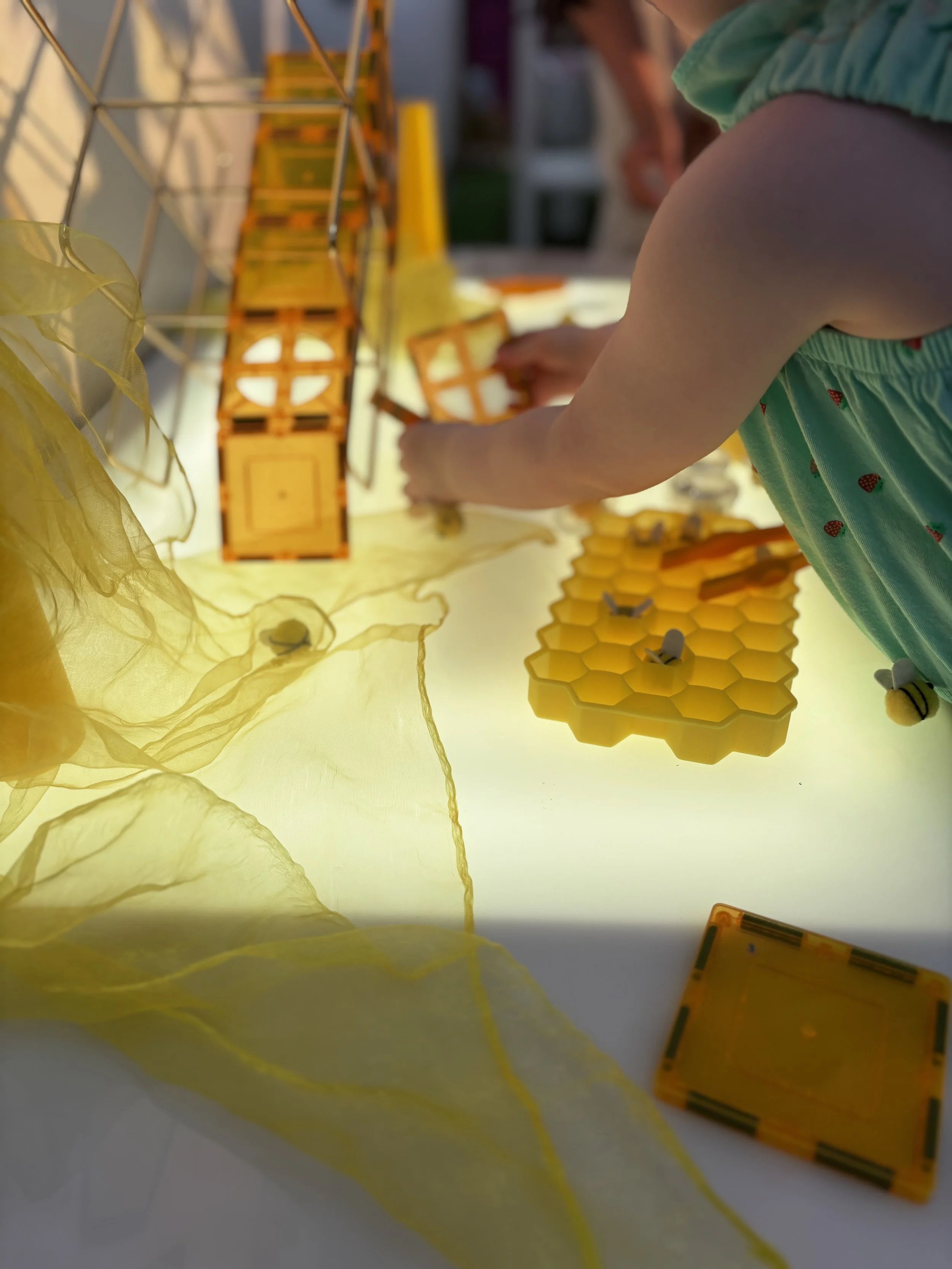 A child is playing with a yellow construction set that resembles a beehive, with honeycomb-shaped pieces and bee figures on top. The child is wearing a sleeveless turquoise dress with small red strawberry patterns and is focused on assembling the pieces on a white surface illuminated by sunlight.