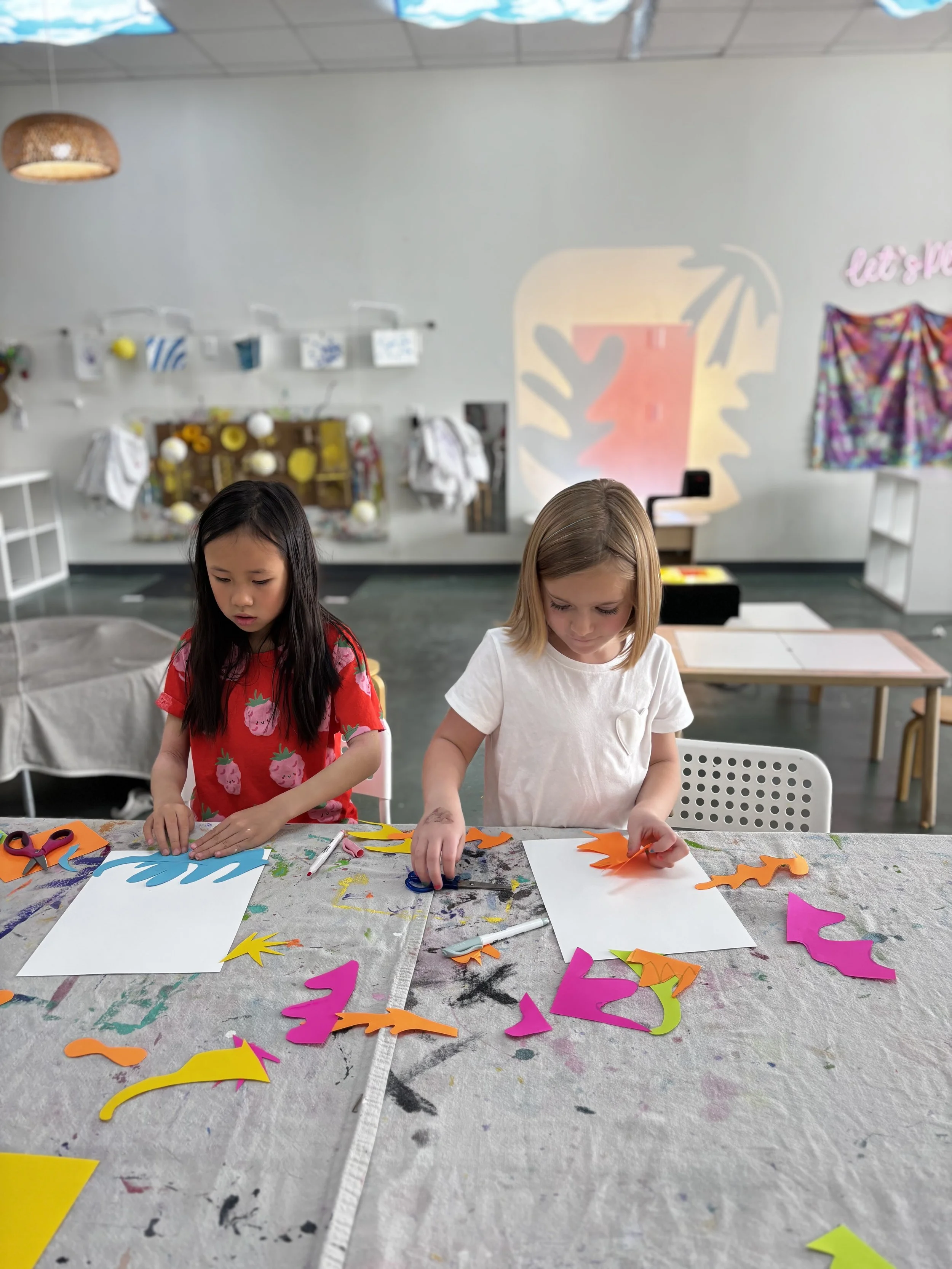 Two young girls working on a craft project at a table with colorful paper cutouts, scissors, and markers in an art classroom. There are art supplies and decorations on the wall in the background.