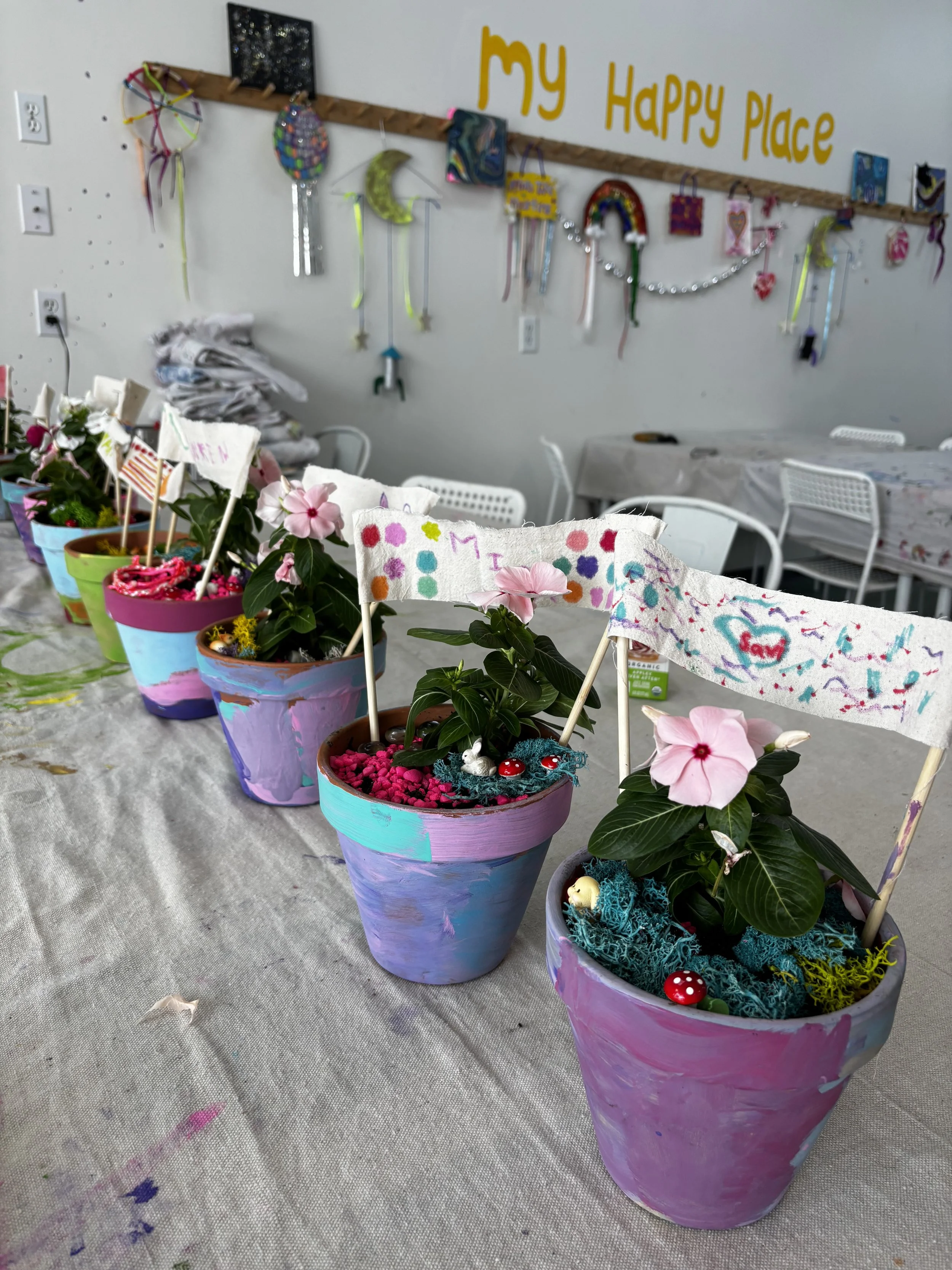 Colorful painted flower pots with blooming flowers and craft flags on a table, with a decorated wall that reads 'My Happy Place' in the background.