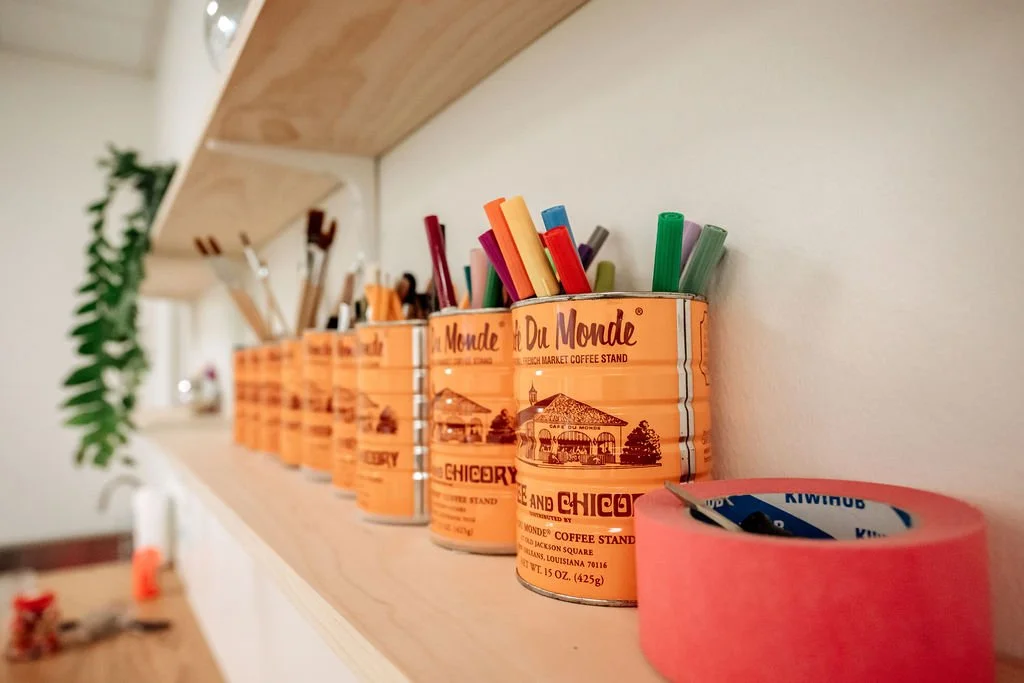Six orange cans used as pen holders on a white shelf, filled with colorful pens and markers, with a roll of pink tape nearby.
