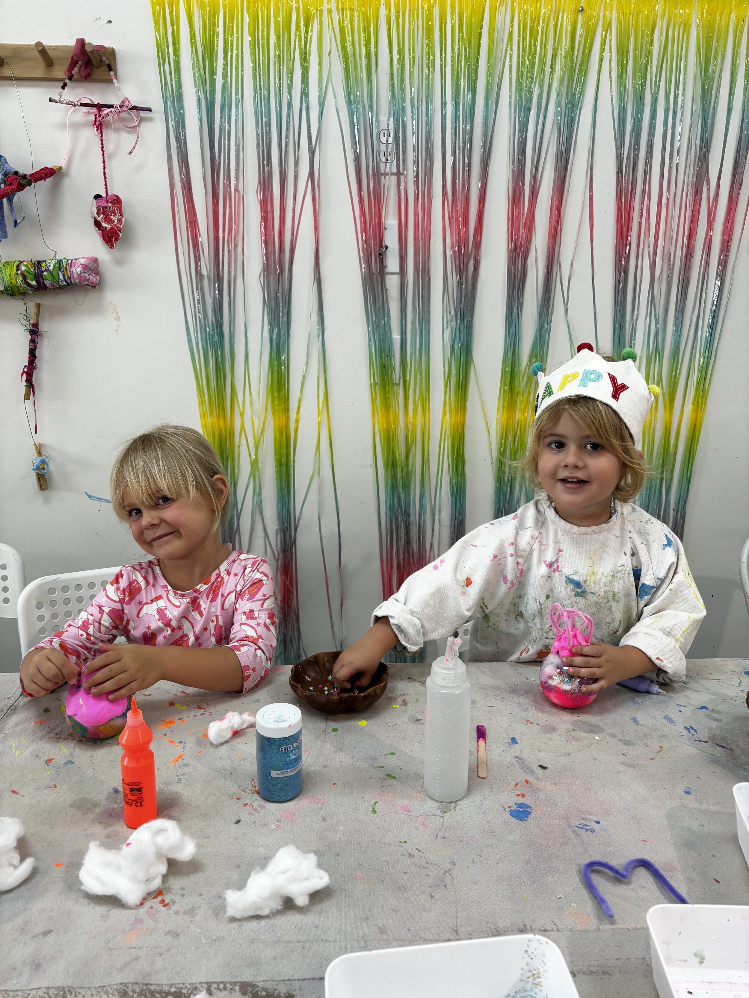 Two young girls sit at a table covered with art supplies, making slime. The girl on the left has blonde hair, is wearing a pink and white patterned shirt, and is holding a pink slime ball. The girl on the right has blonde hair, is wearing a white shirt and a white birthday crown decorated with the word 'HAPPY', and is also holding a pink slime. The background features colorful hanging streamers in yellow, green, and pink.