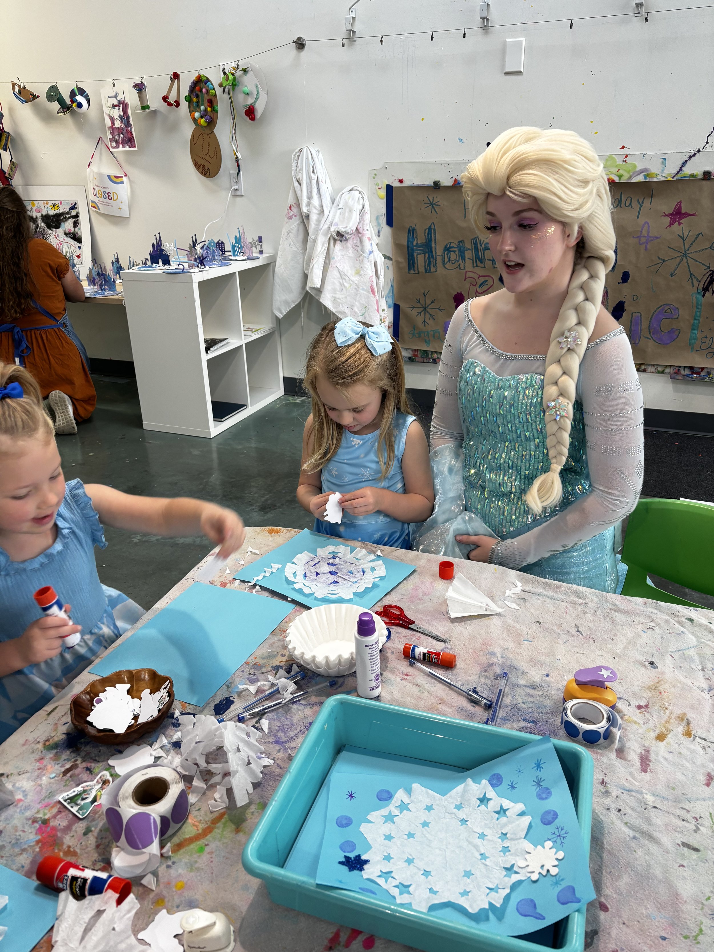 A woman dressed as Elsa from Frozen is supervising children during a holiday-themed arts and crafts activity in a classroom.