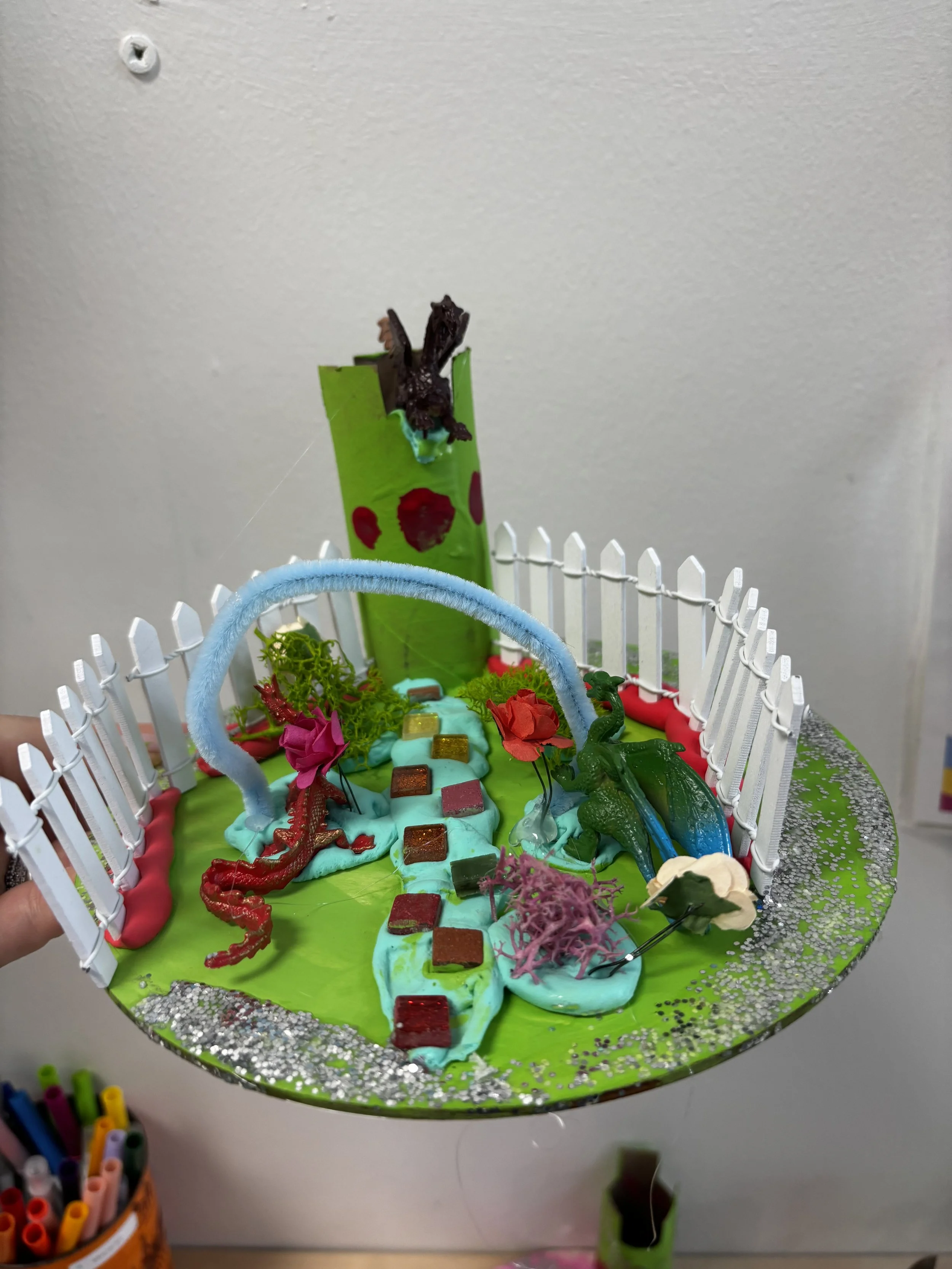 A colorful, miniature homemade science-themed diorama featuring a small volcano with a lava explosion, plastic dinosaurs, a winding path with small colored tiles, greeen foliage, a white picket fence, and decorative glitter on a green base.