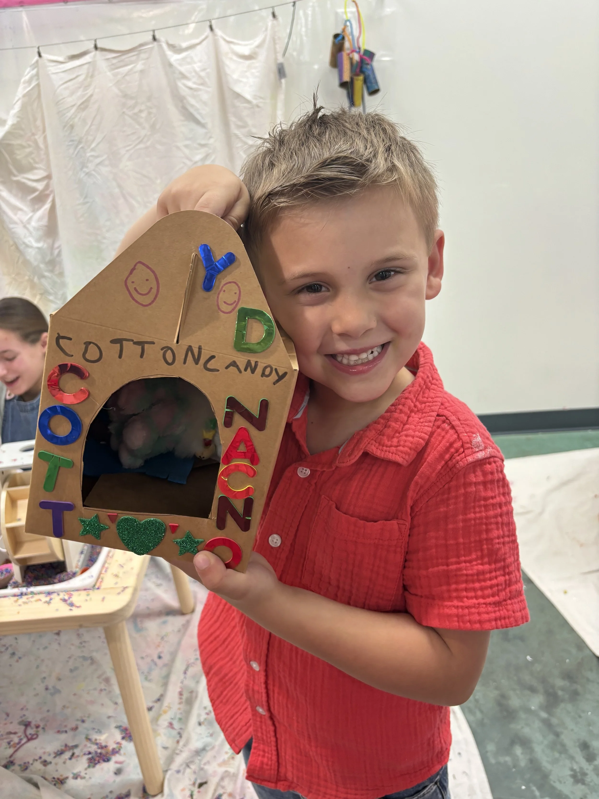 A young boy wearing a red shirt is smiling and holding a cardboard box decorated with colorful letters and shapes, with the words 'cotton candy' written on it, in what appears to be a classroom or craft area.