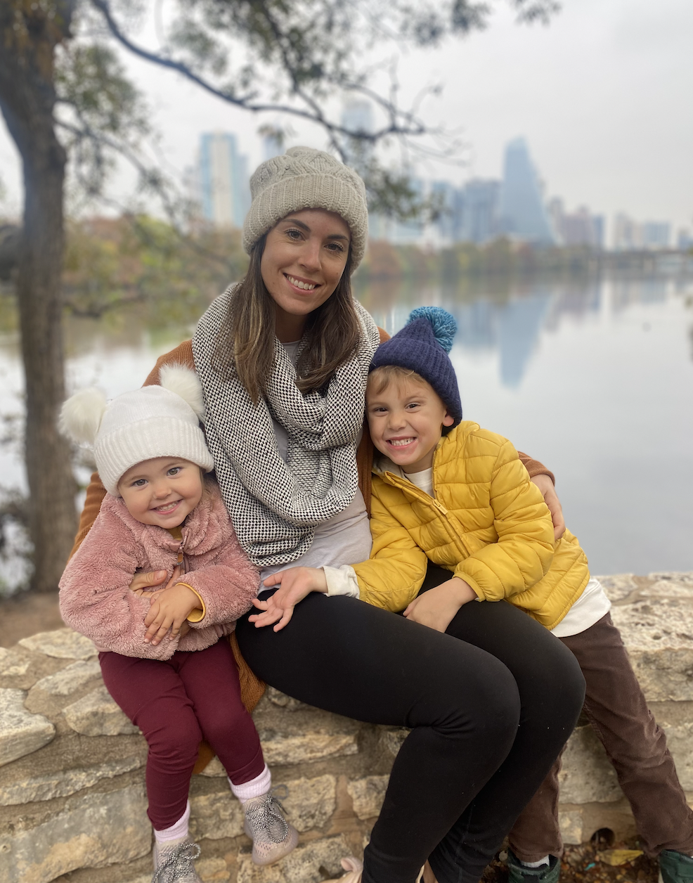 A woman and two children sitting on a stone ledge outdoors near a lake with a city skyline in the background, dressed warmly in winter clothing.