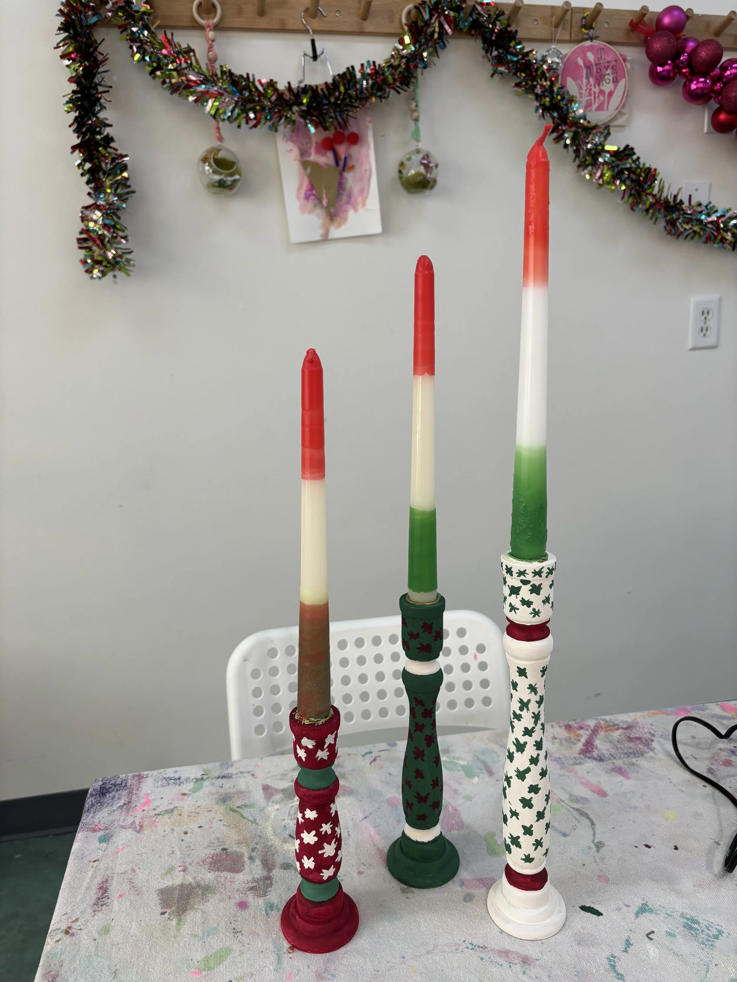 Three tall candles painted in red, white, and green with festive patterned holders, placed on a table covered with a paint-splattered cloth. In the background, holiday garlands and ornaments are hanging on a wall.