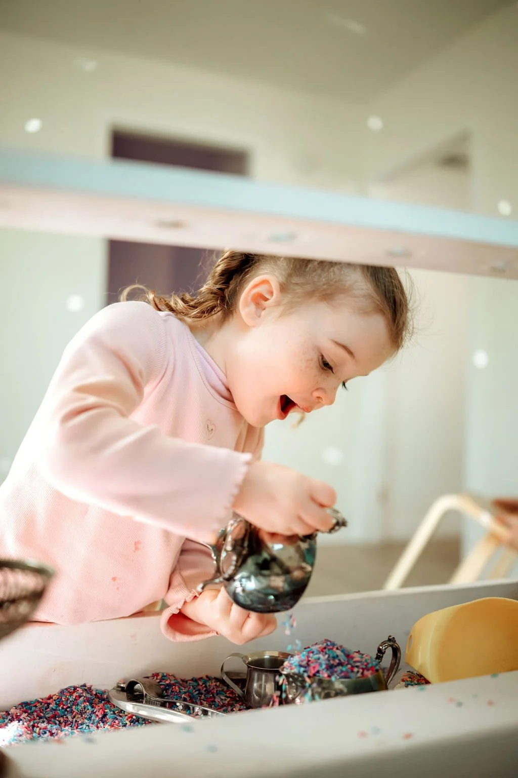 A child is playing with a yellow construction set that resembles a beehive, with honeycomb-shaped pieces and bee figures on top. The child is wearing a sleeveless turquoise dress with small red strawberry patterns and is focused on assembling the pieces on a white surface illuminated by sunlight.