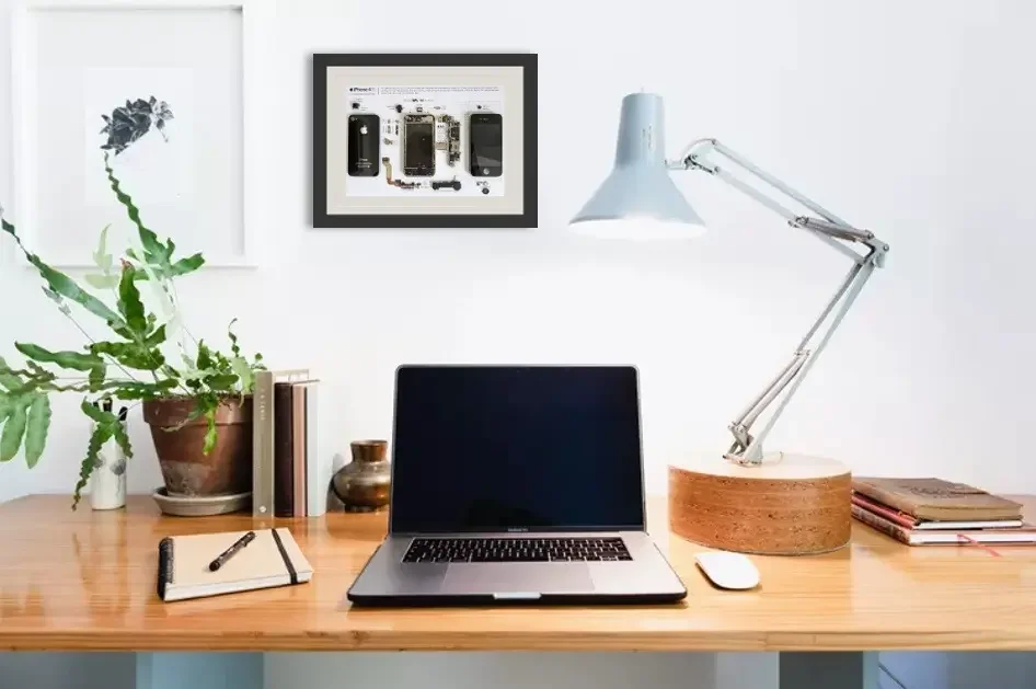 A tidy workspace with a laptop, desk lamp, potted plant, notebooks, and a framed diagram of electronic devices on a white wall.