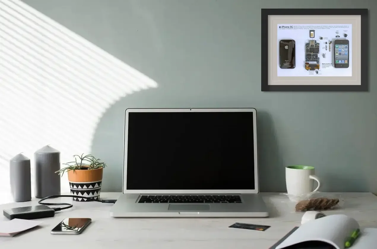 A workspace with a silver laptop, a plant in a black and white pot, a green and white mug, a smartphone, a few documents, two grey candles, a remote, and a framed display of disassembled iPhone parts on a light-colored desk against a light blue wall.