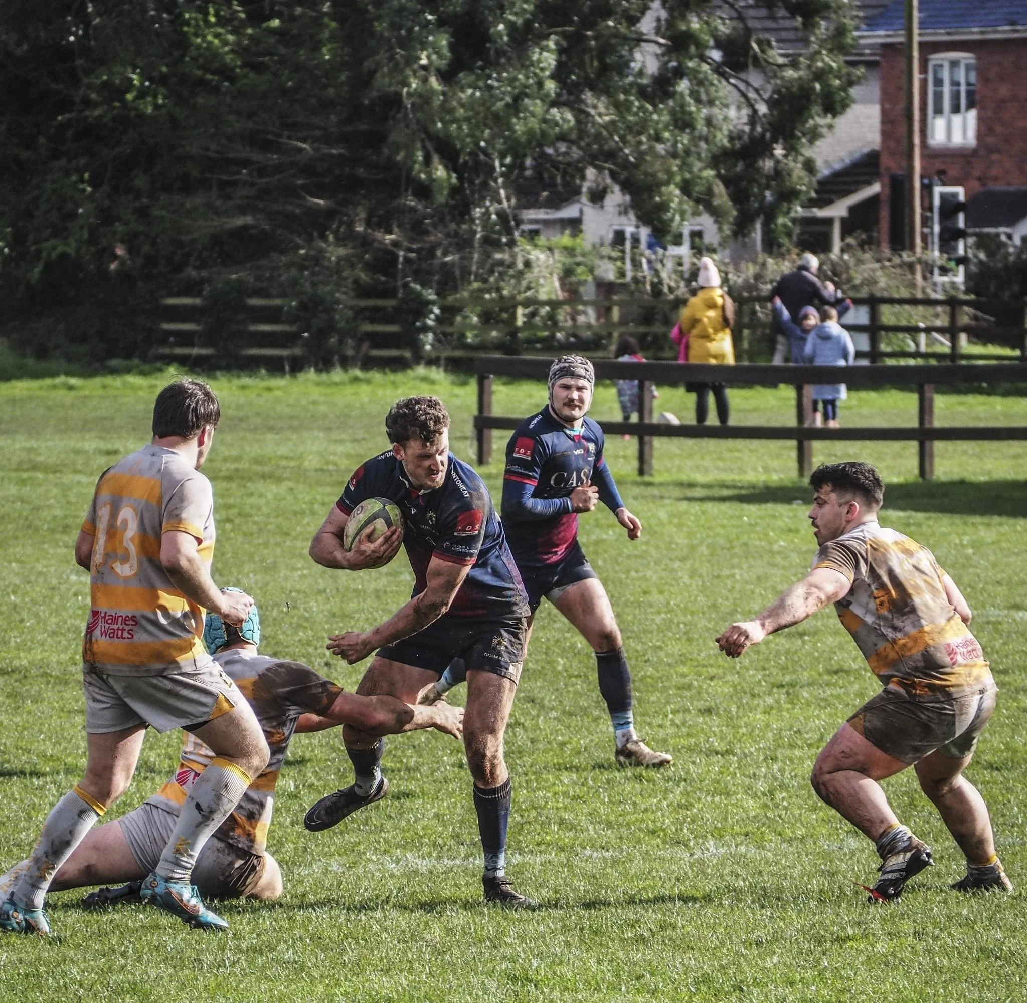 Rugby players in action on a grassy field, with one player running with the ball while others move to block or tag him; muddy uniforms and spectators watching in the background.