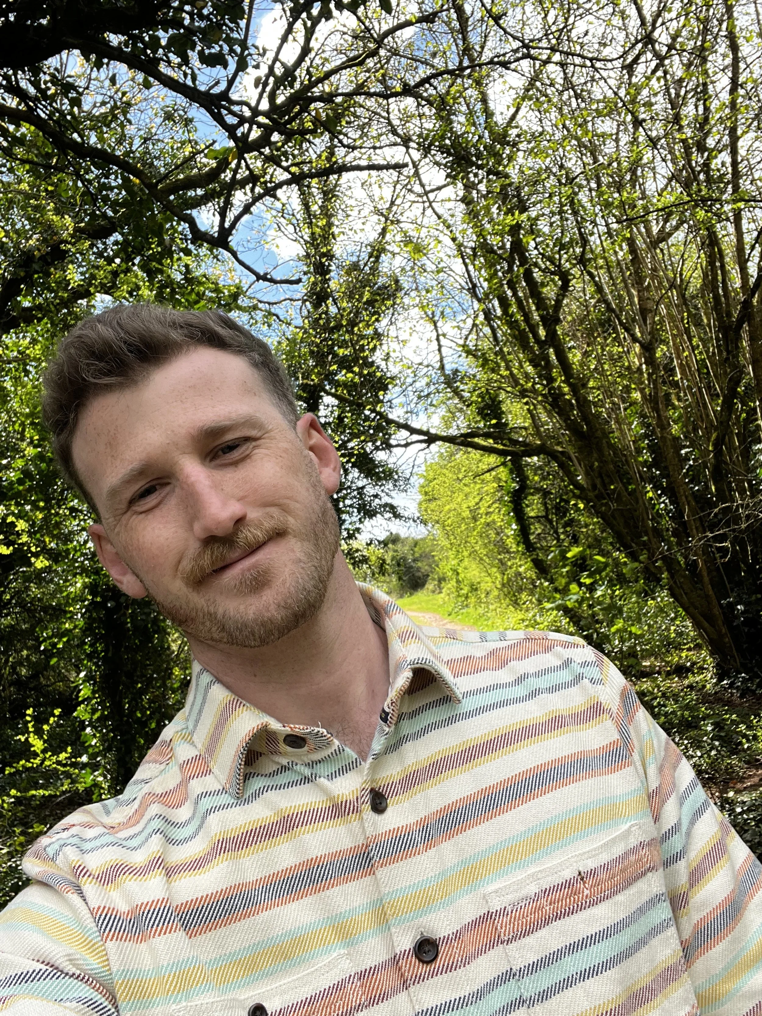 A smiling young man with light skin, a mustache, and short curly brown hair, wearing a colorful striped button-up shirt, standing on a nature trail surrounded by green trees and bushes with blue sky visible overhead.