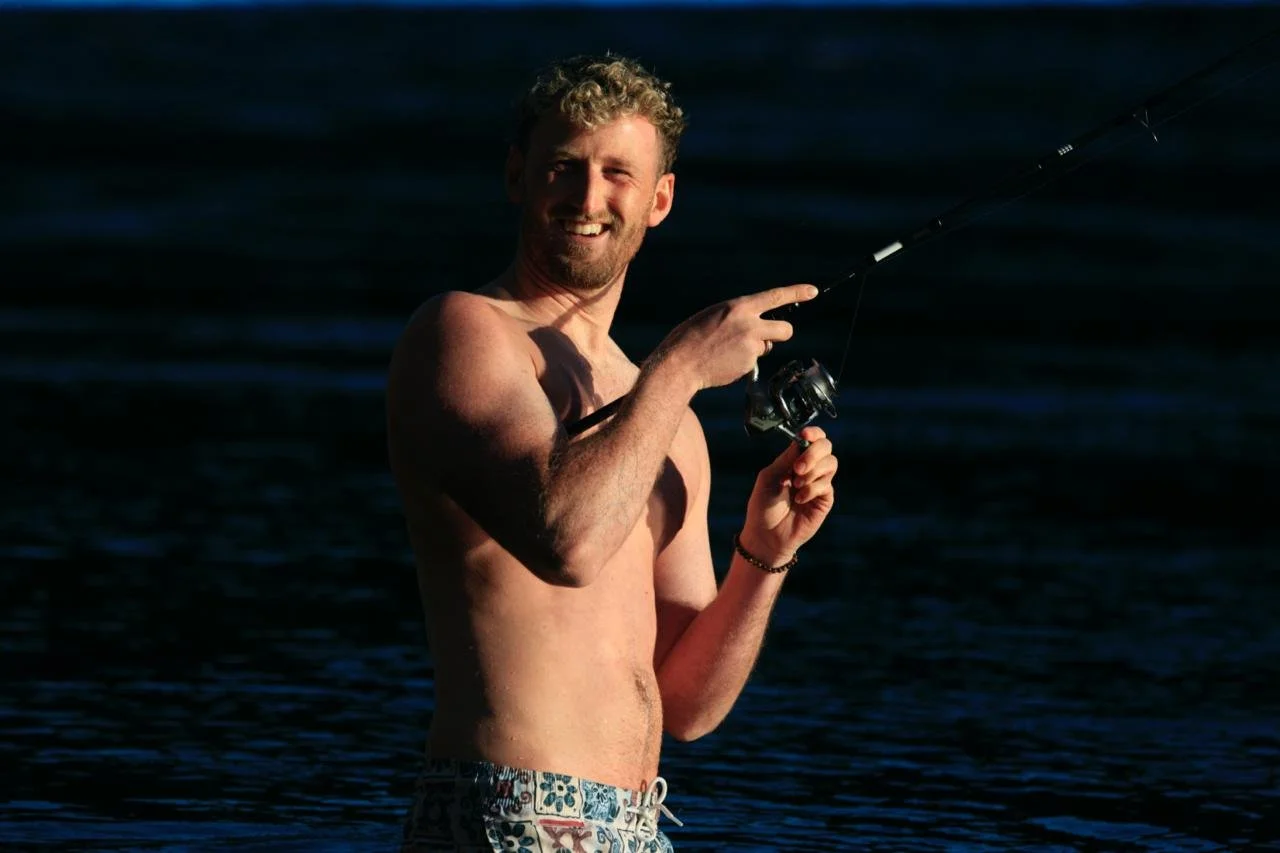 A shirtless man with curly hair smiling while fishing in a body of water during daylight.