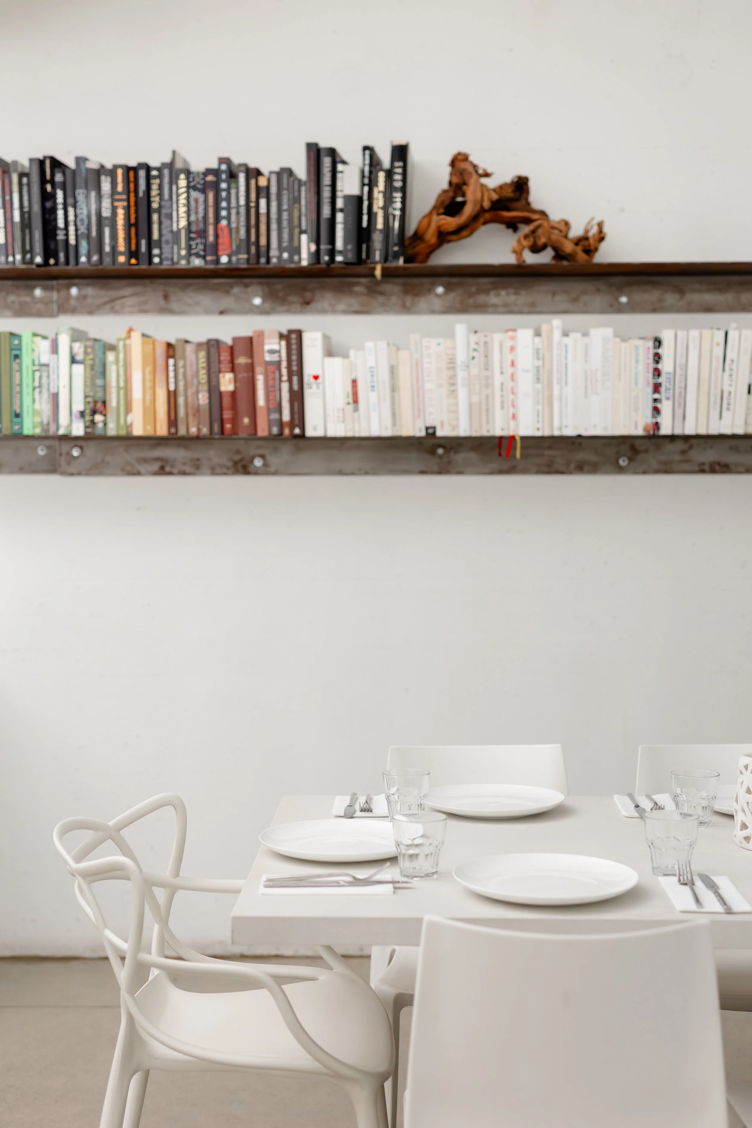 A modern dining table set with white plates, glasses, and cutlery, in front of a white wall with a bookshelf filled with books.