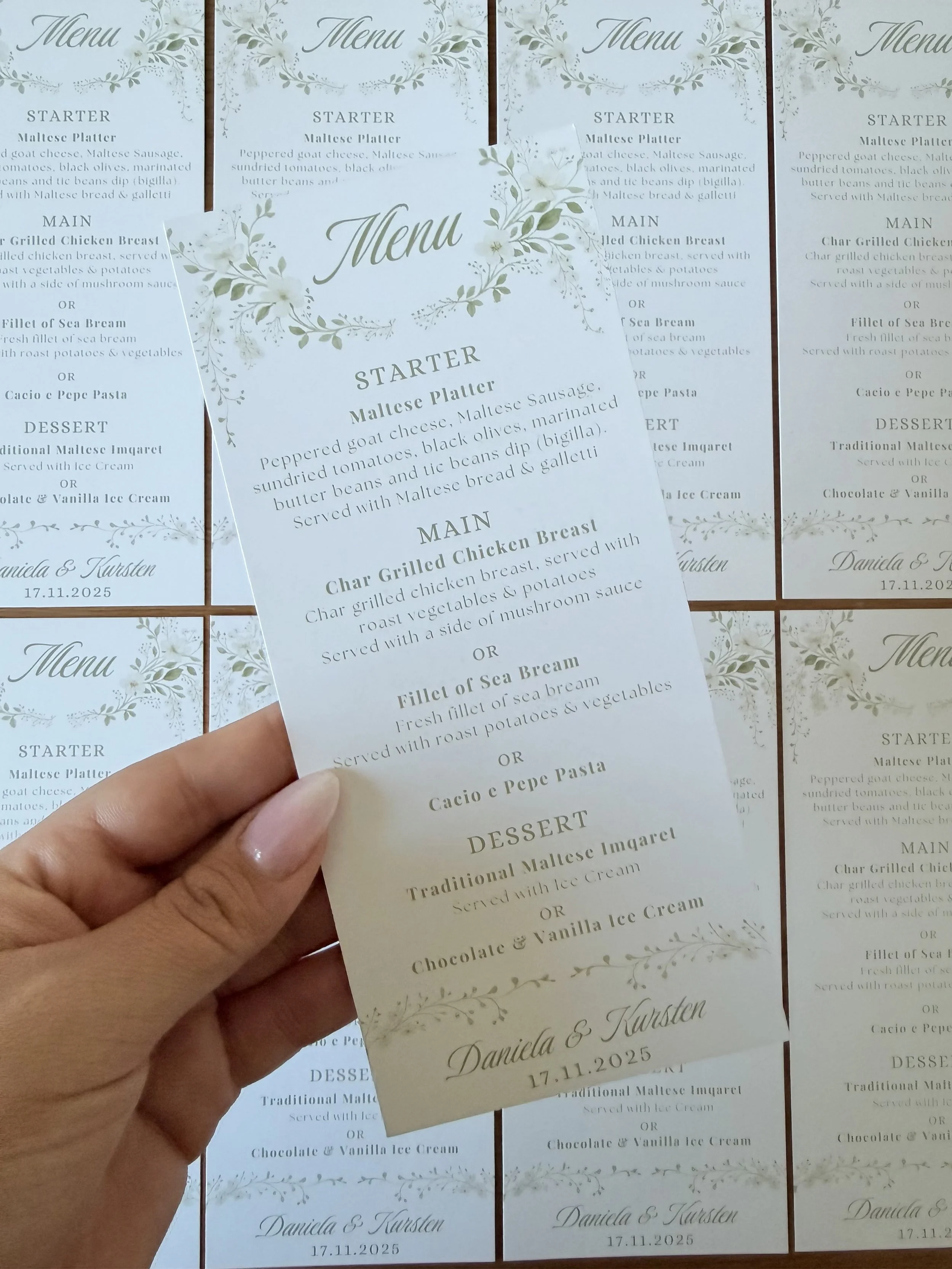 Close-up of a wedding menu card with floral design, listing starter, main courses, and dessert options, held over similar menus on a table.