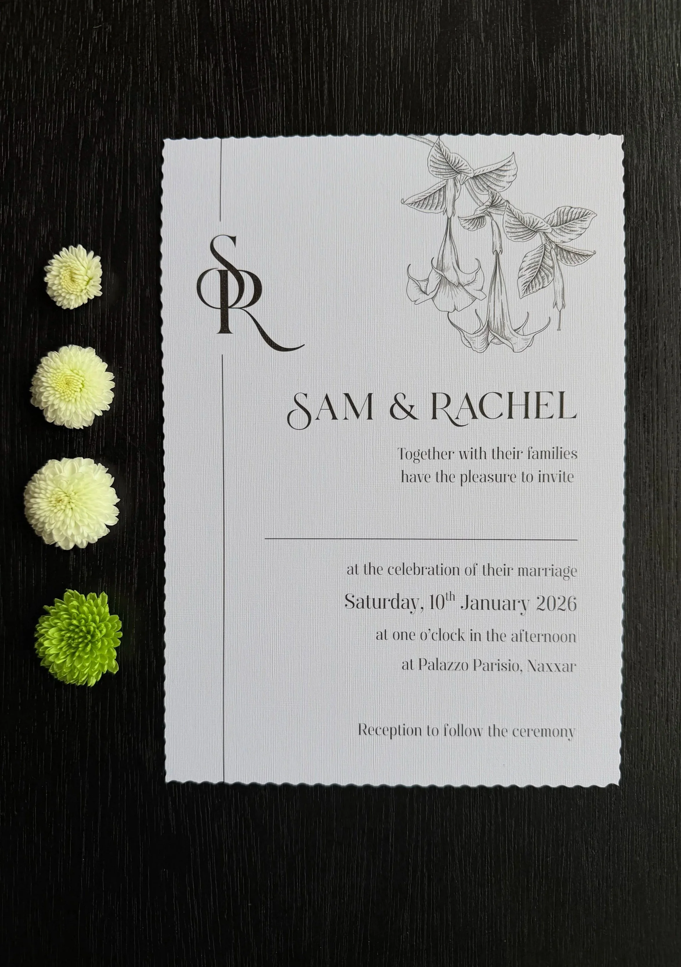 Wedding invitation card for Sam and Rachel, with floral illustrations, placed on a black surface with four white and green flowers to the left.