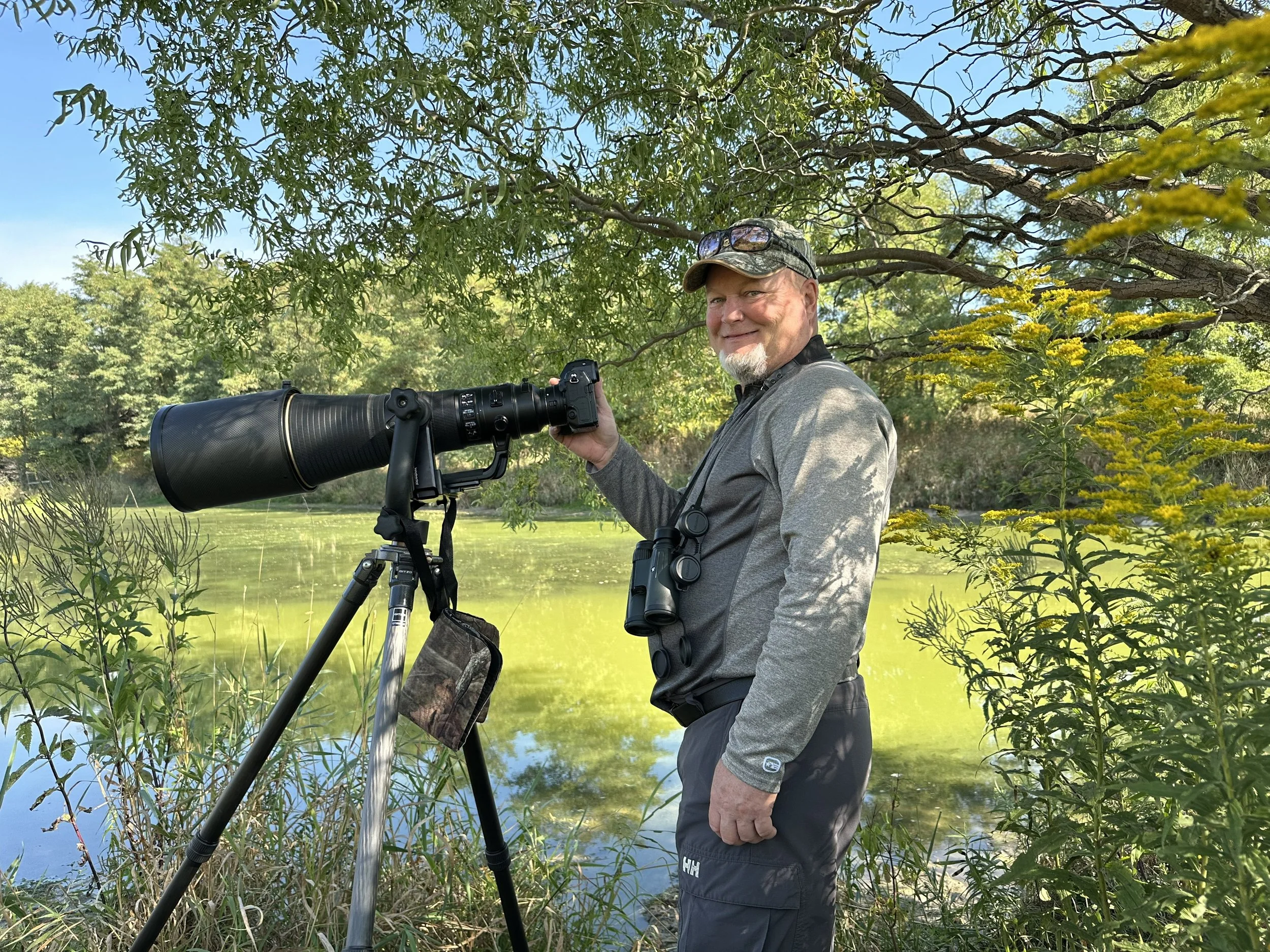 Terry Parker standing outdoors beside a pond in Oxford County with a large camera on a tripod. He is capturing images of South Western Ontario birds at the family farm.