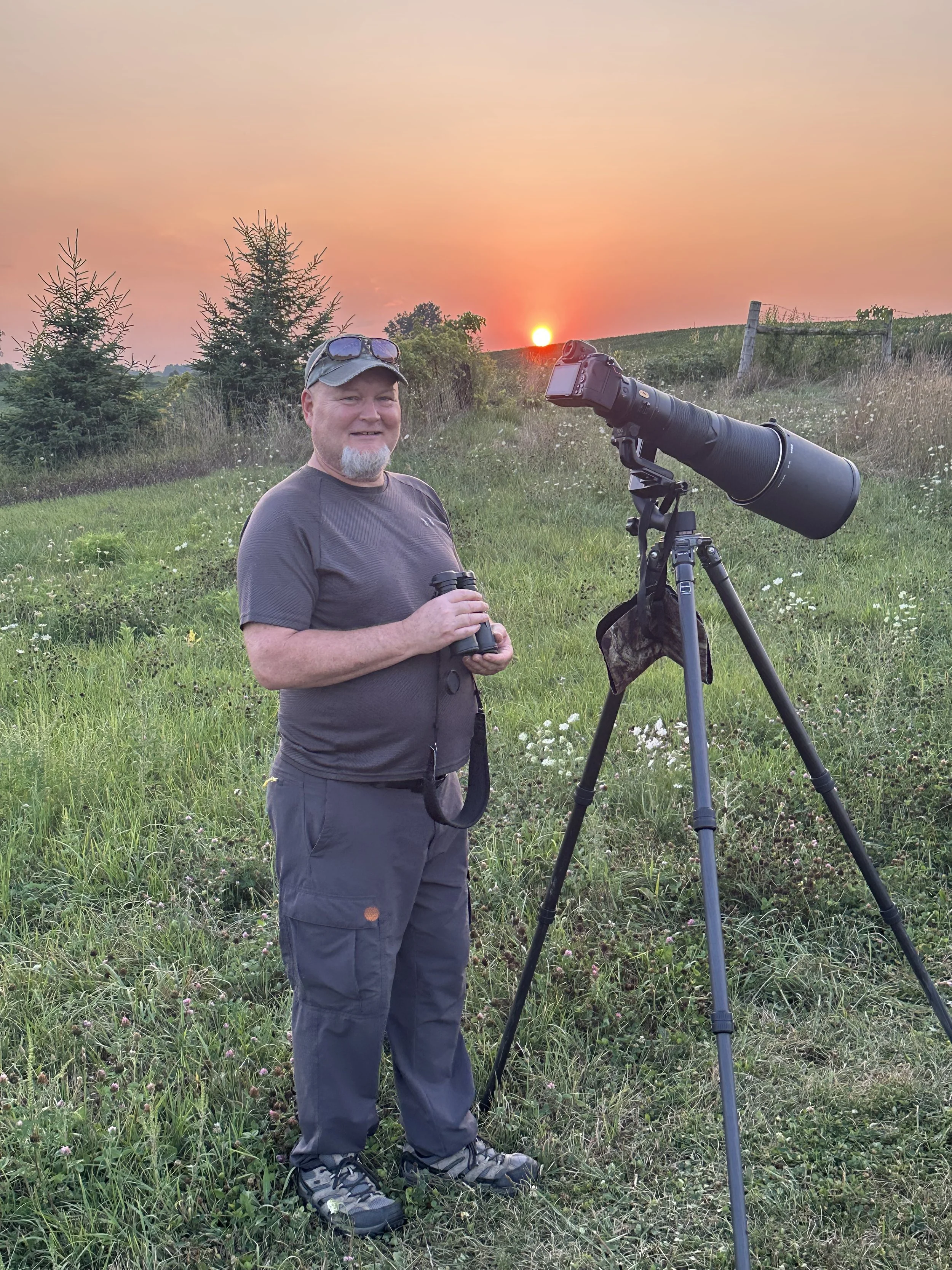 A man standing outdoors at sunset with a camera telescope on a tripod, holding binoculars, surrounded by green grass and trees.