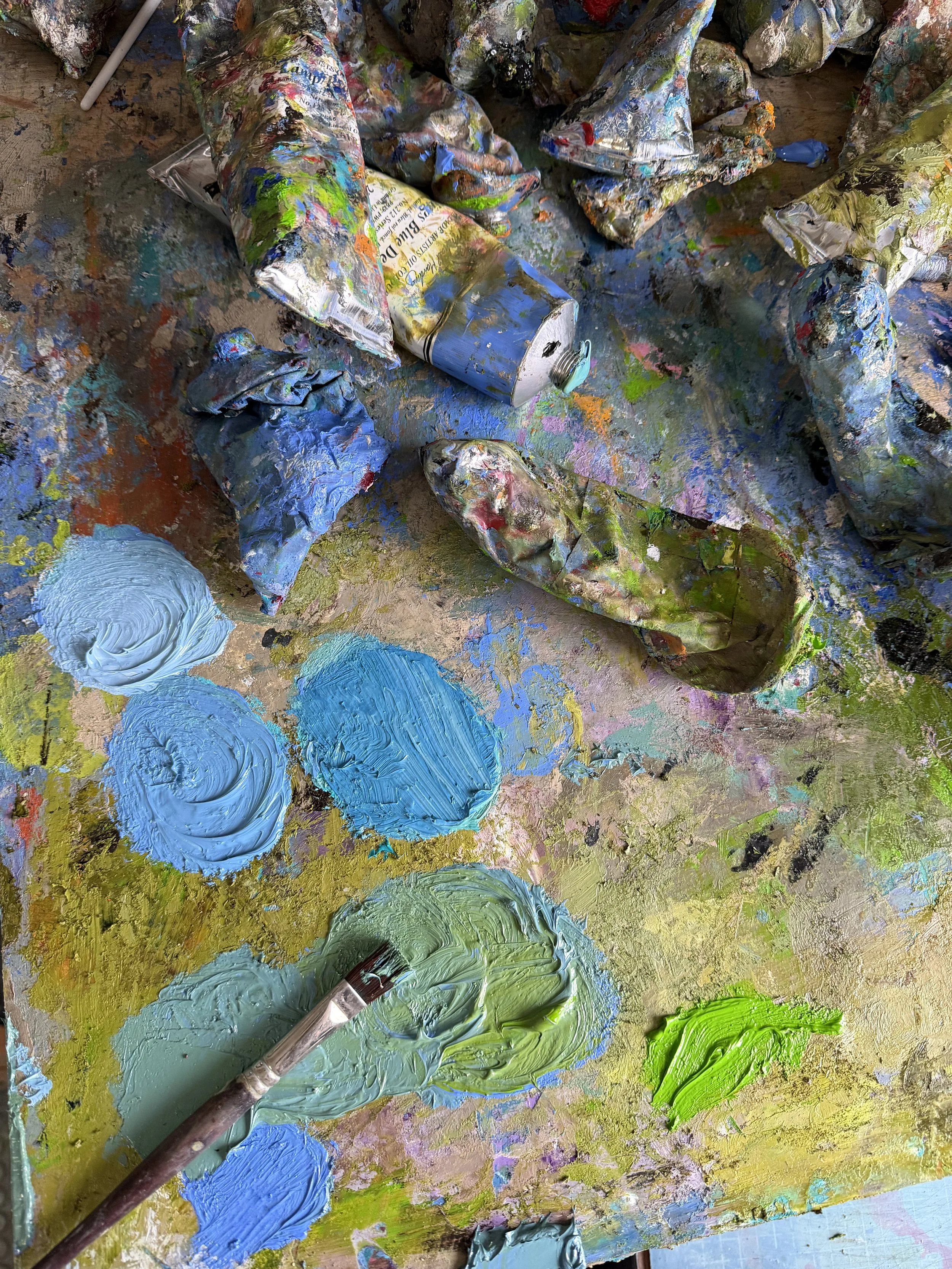 A cluttered painting workspace with messy, crumpled paint tubes, paint smears, and blobs of blue, green, and yellow oil paints on a paint-covered surface.