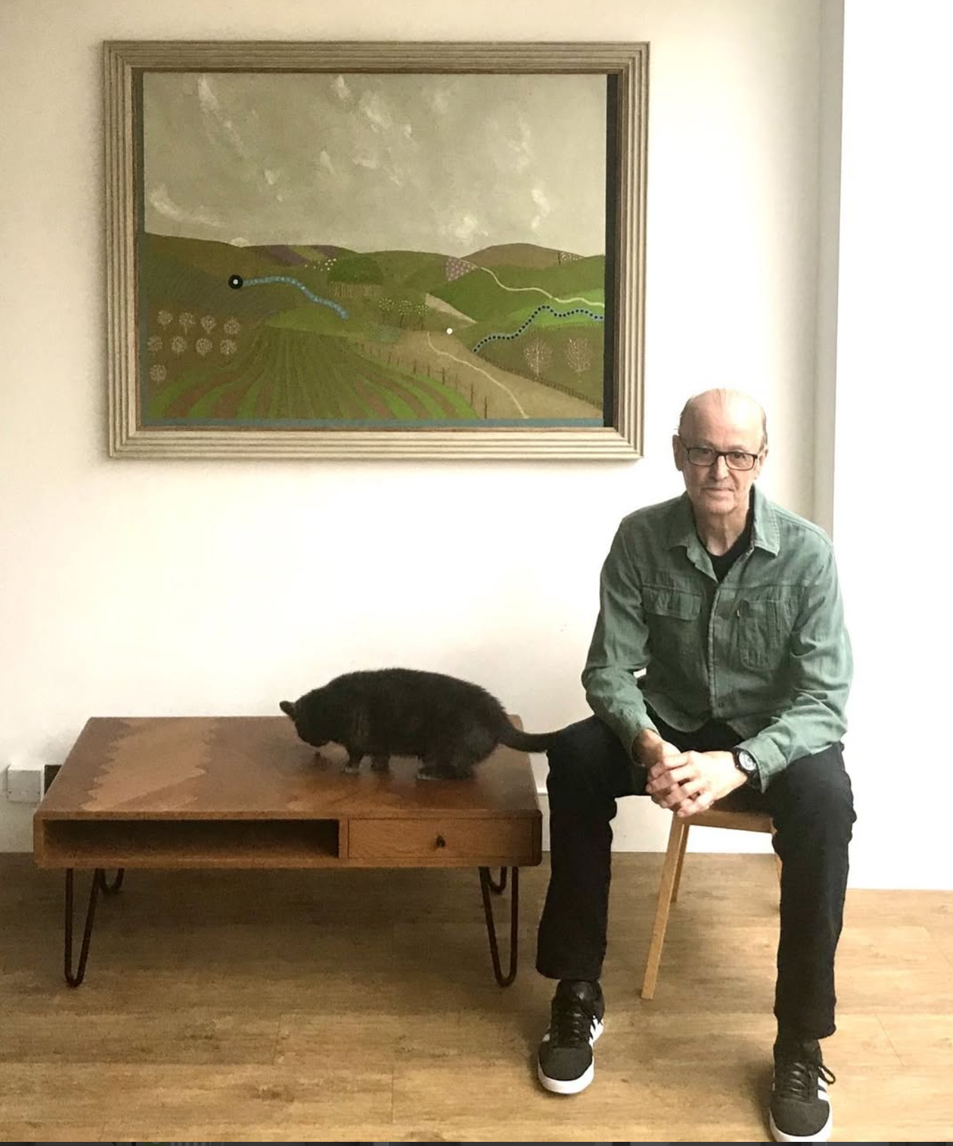 A man with glasses in a green shirt and black pants sitting on a wooden chair next to a small black animal on a wooden table. There is a man-made framed landscape painting of green fields and rolling hills hanging on the wall behind them.