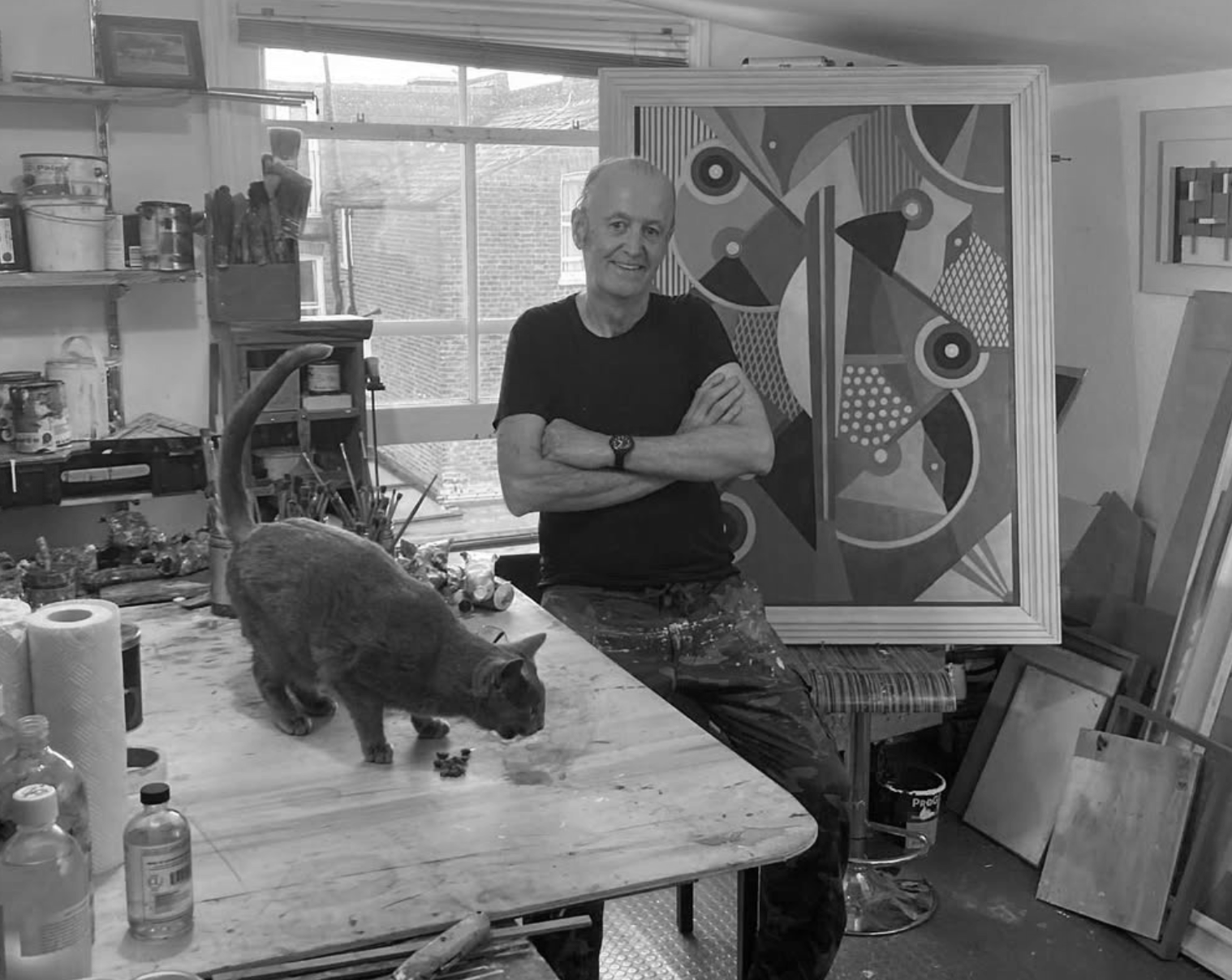 An artist's studio with a man standing next to a large abstract painting. The studio has art supplies and canvases, with a cat on the table.