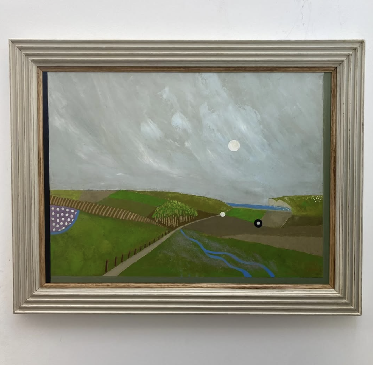 A landscape painting depicting a dirt path, green hills, a small stream, and a cloudy sky with a visible moon, framed in a silver wooden frame.