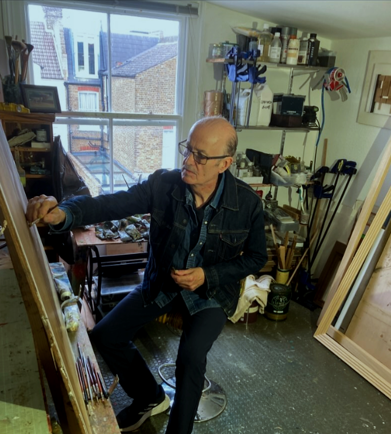 A man sitting on a stool in a cluttered art studio, painting on a canvas. The studio has a large window with a view of neighboring brick buildings and is filled with art supplies, frames, and tools.