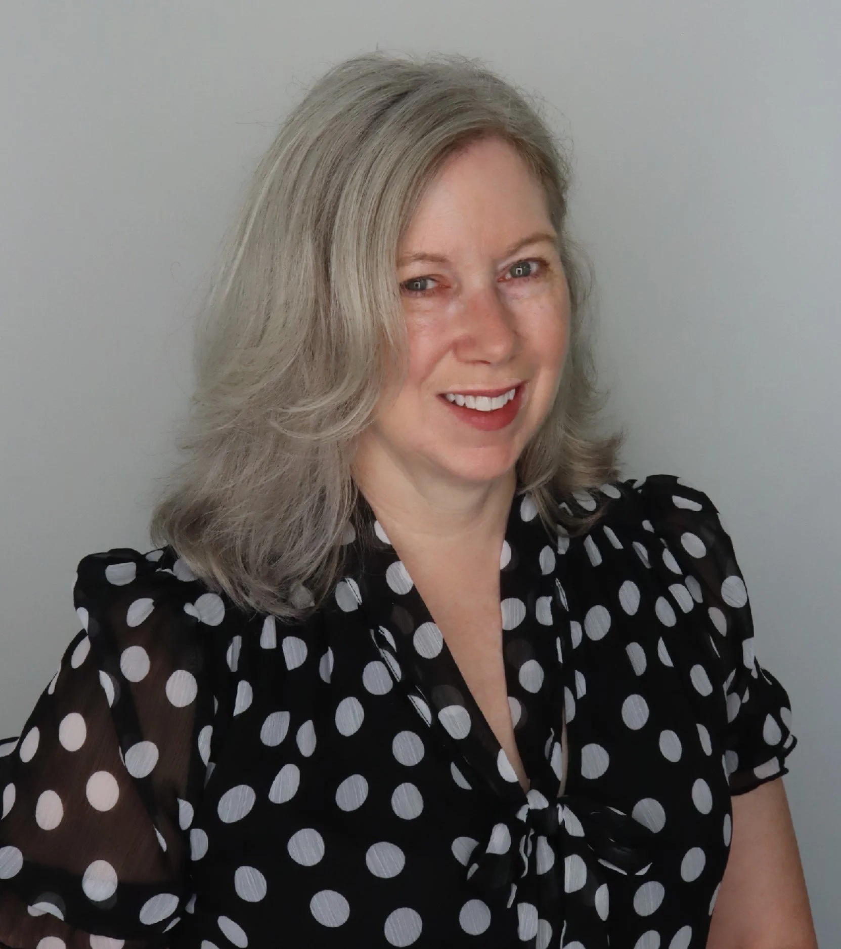 A woman with shoulder-length gray hair smiling, wearing a black blouse with white polka dots, standing against a plain light-colored background.
