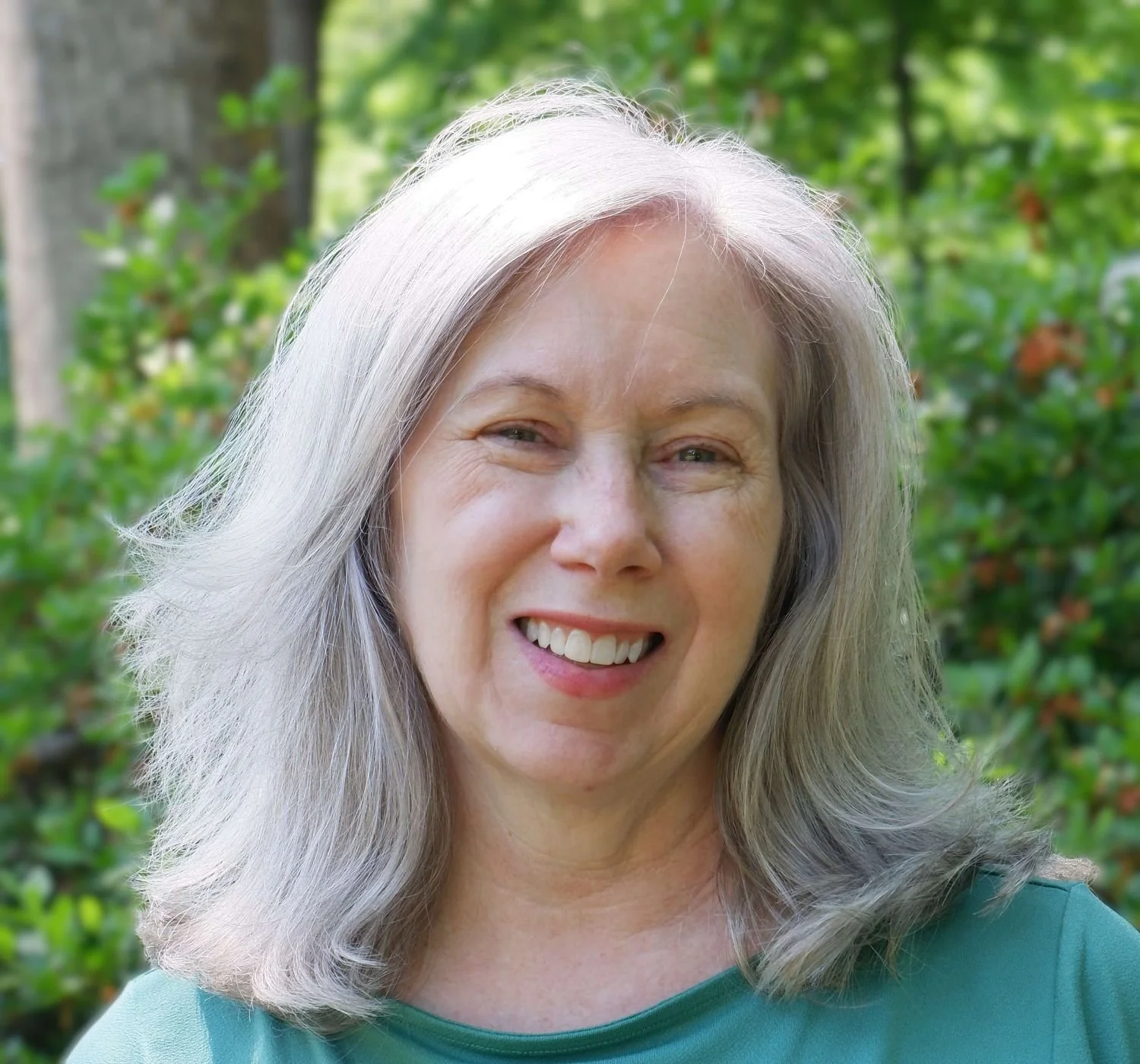 Portrait of a smiling middle-aged woman with long gray hair, wearing a green shirt, outdoors among green foliage.