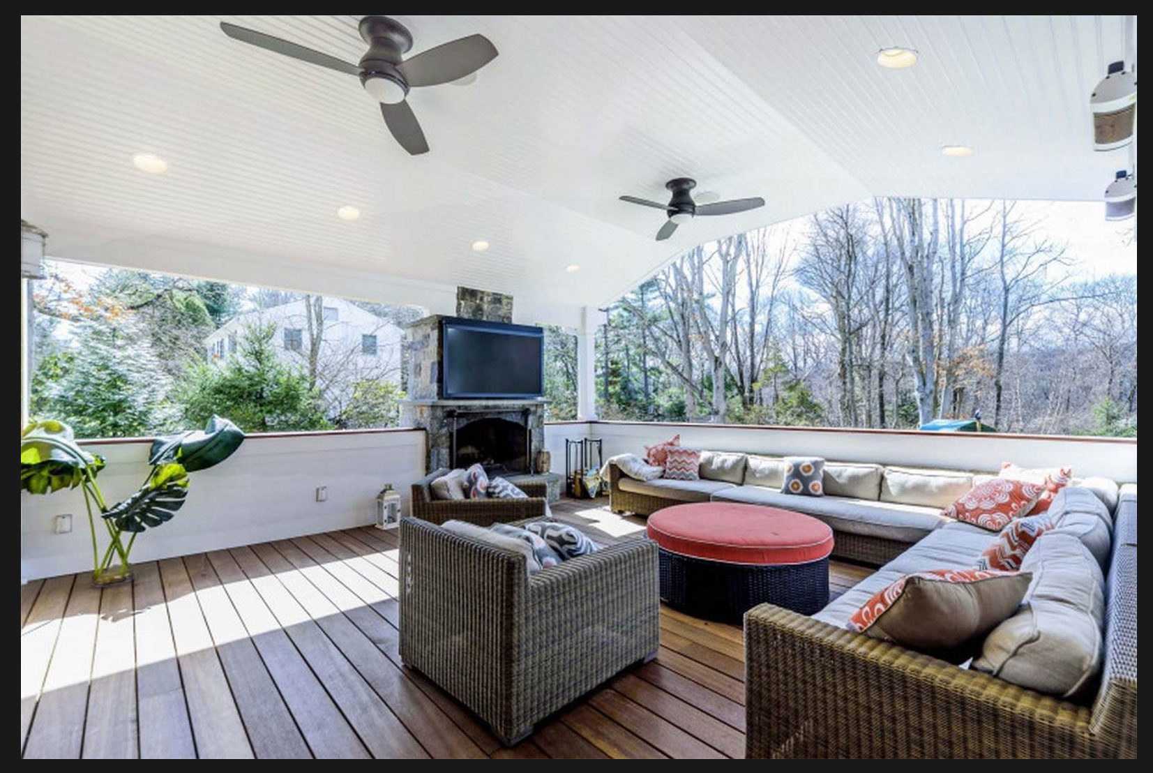 Covered outdoor patio with ceiling fans, sectional sofa with decorative pillows, round ottoman, armchairs, a fireplace with a TV mounted above, and trees in the background.