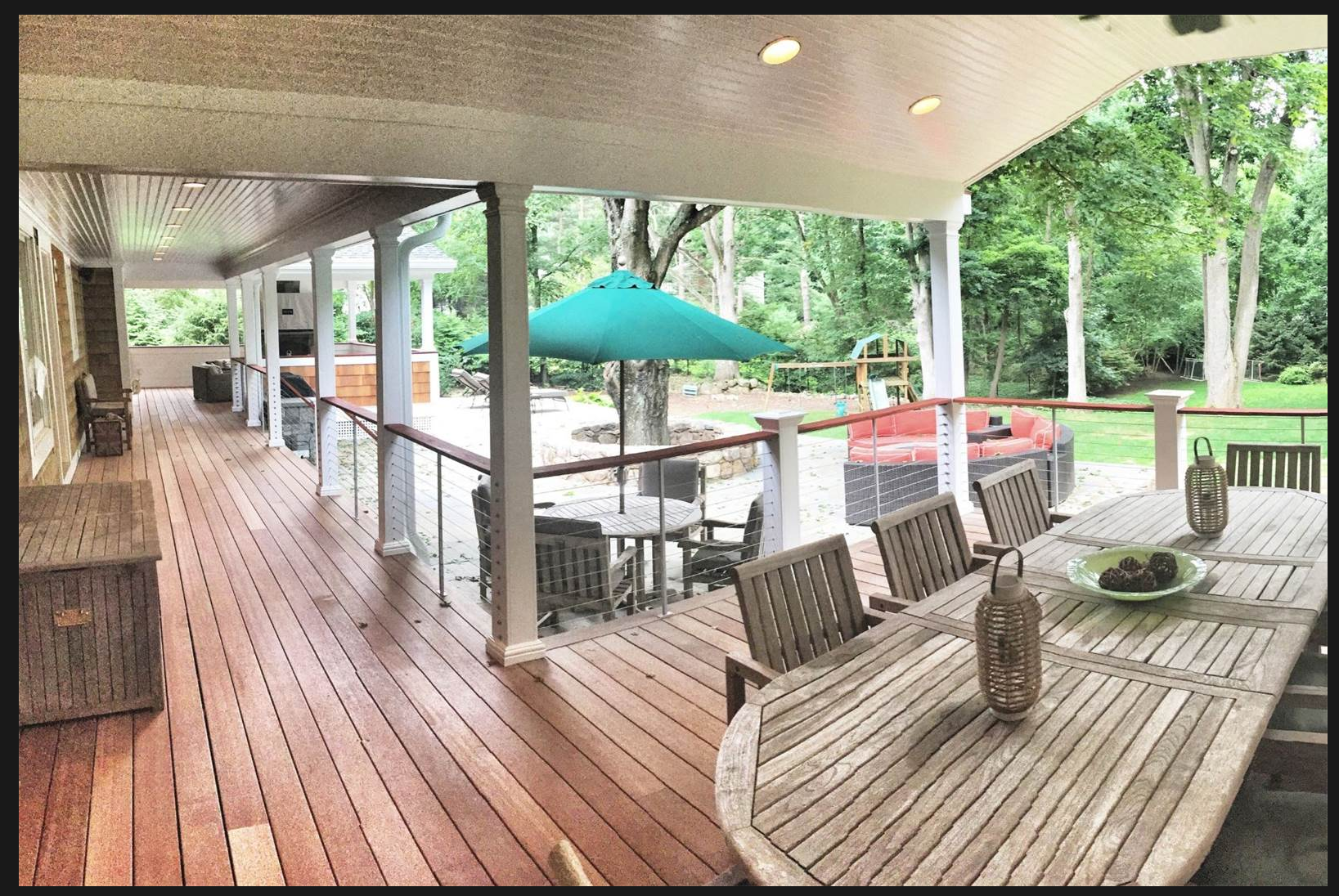 Covered backyard patio with wooden deck, outdoor furniture including a dining table with chairs, a small round table with chairs, and a sofa under a large green umbrella, surrounded by a yard with trees and greenery.